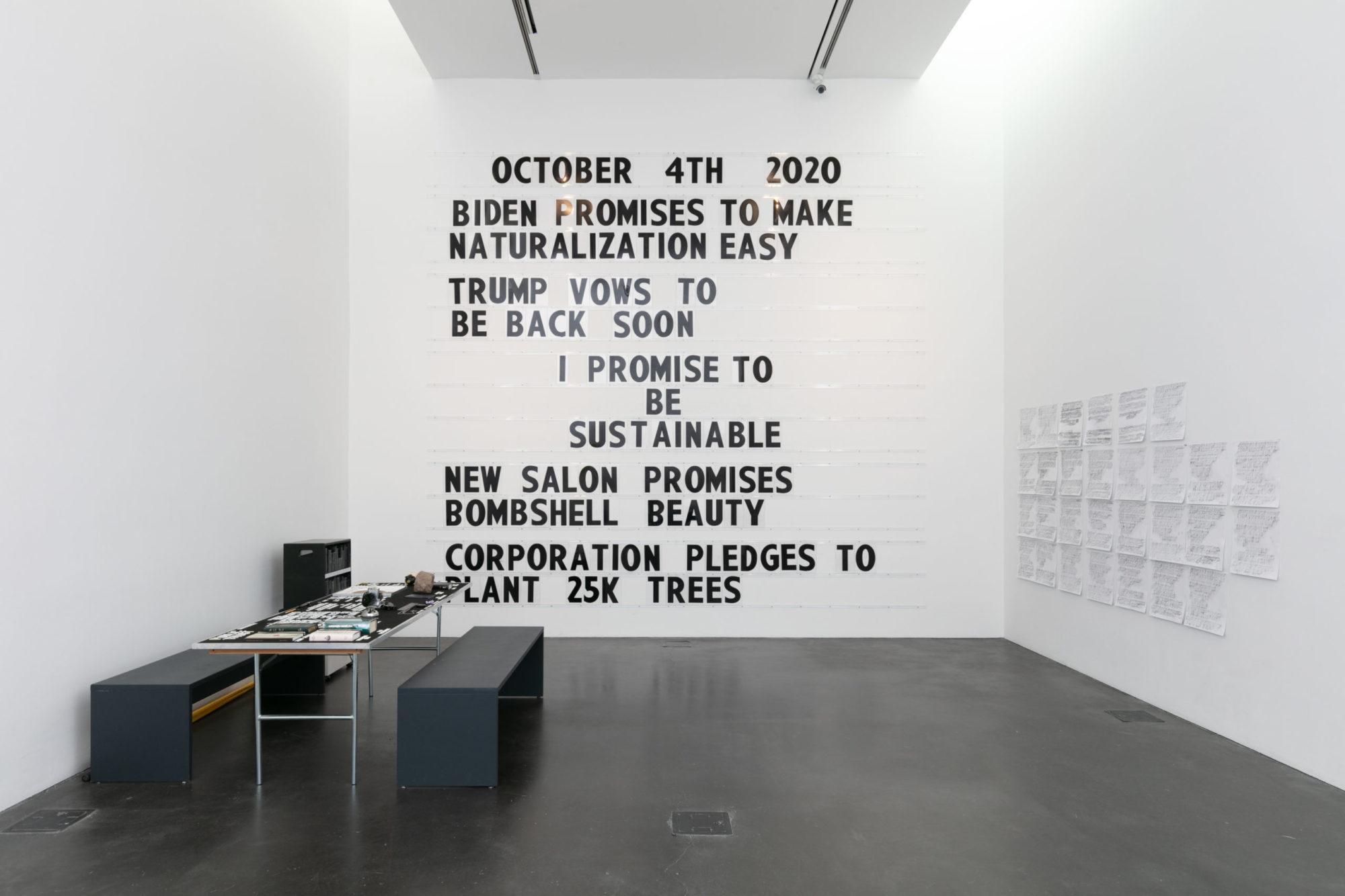 Marquee with black letters on white gallery wall spelling out five promises made by policy makers and private citizens, adjacent wall covered in grid of paper, desk stands against wall