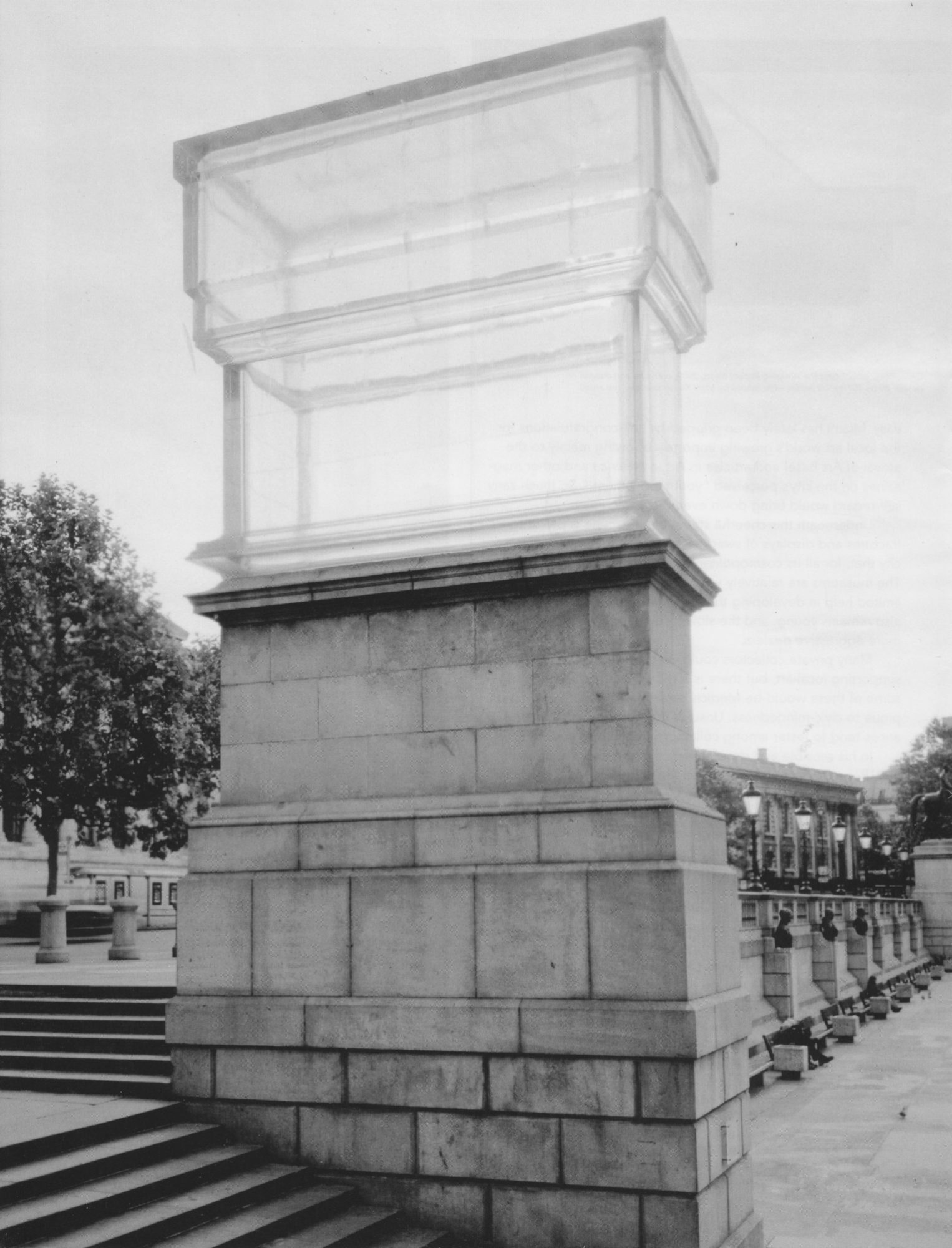 A transparent sculpture of a two-leveled wardrobe atop a stone column. It looks like a drawer or dresser from the behind, with the bottom half being a smaller portion. Around the sculpture is an outdoor atrium, a walkway and trees behind it, with stairs leading up to it.