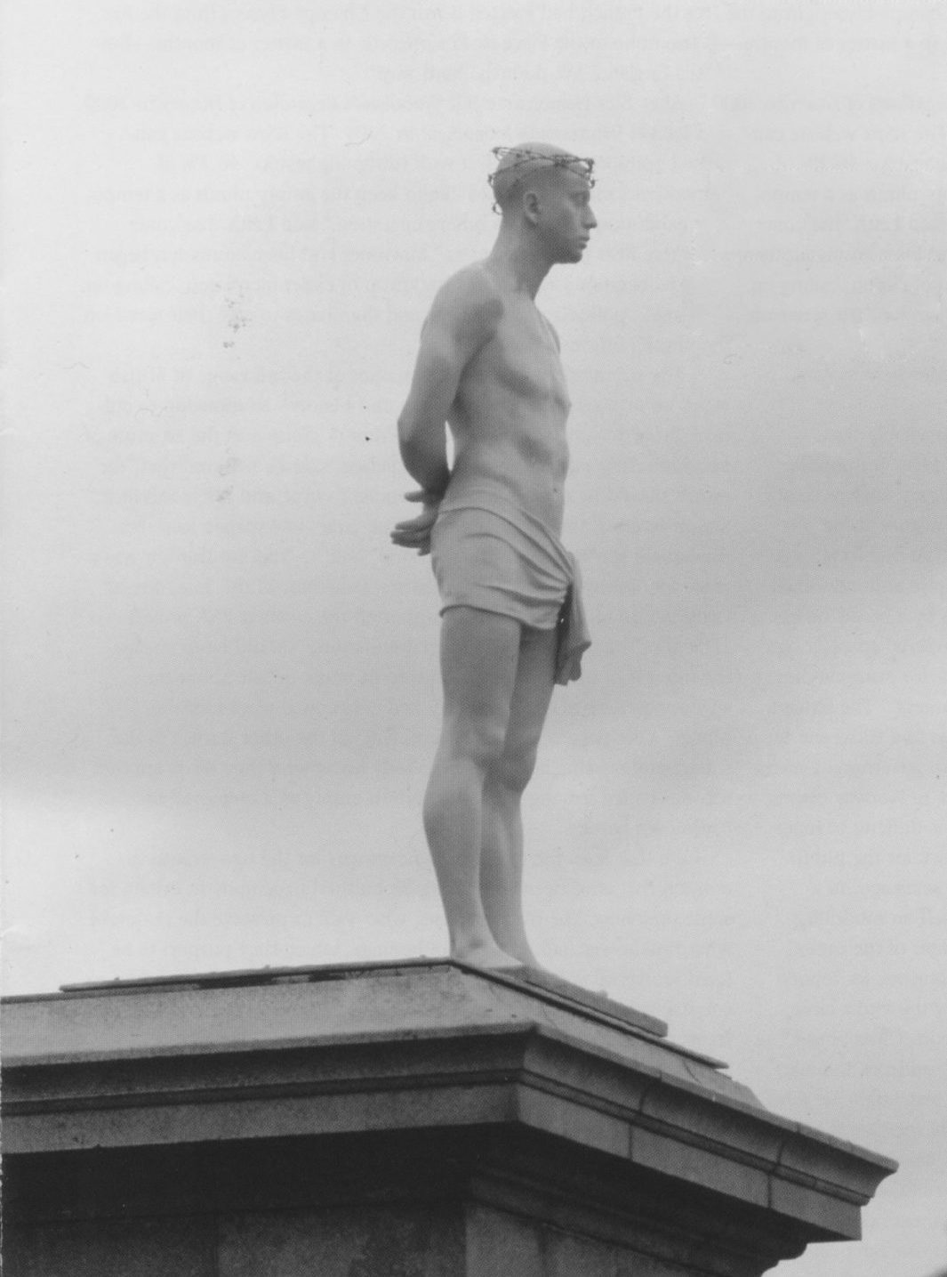 A lone sculpture of a single man painted white standing atop a large column and standing faced to the right. His hands are behind his back. The man wears only a white cloth around his pelvis, and a hair adornment. Behind him are clouded skies.
