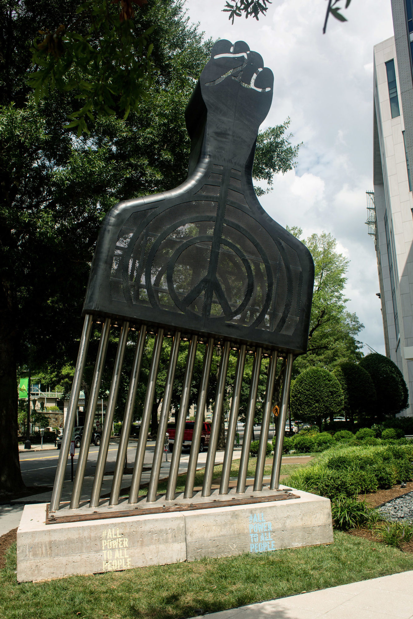 A large sculpture of a hair pick with a fist pointing outward on its handle and a peace sign in the center. The pick is stuck sideways into a plot of concrete over grass.