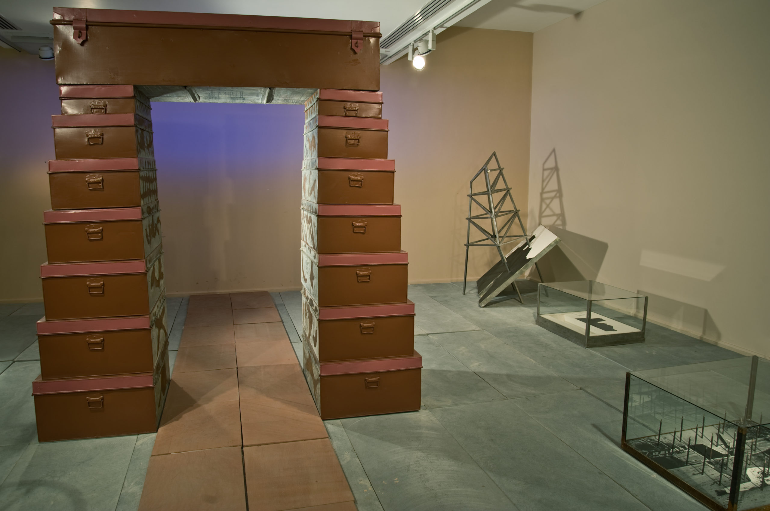 Close up of a brown altar, which is made of stacked cabinets or drawers. Each layer of cabinets are smaller than those underneath them. At the top, there's one long cabinet which creates the roof of the altar. To the right there are several smaller pieces of sculpture placed in glass tanks, with another sculpture in the corner which looks like a gray telephone line.