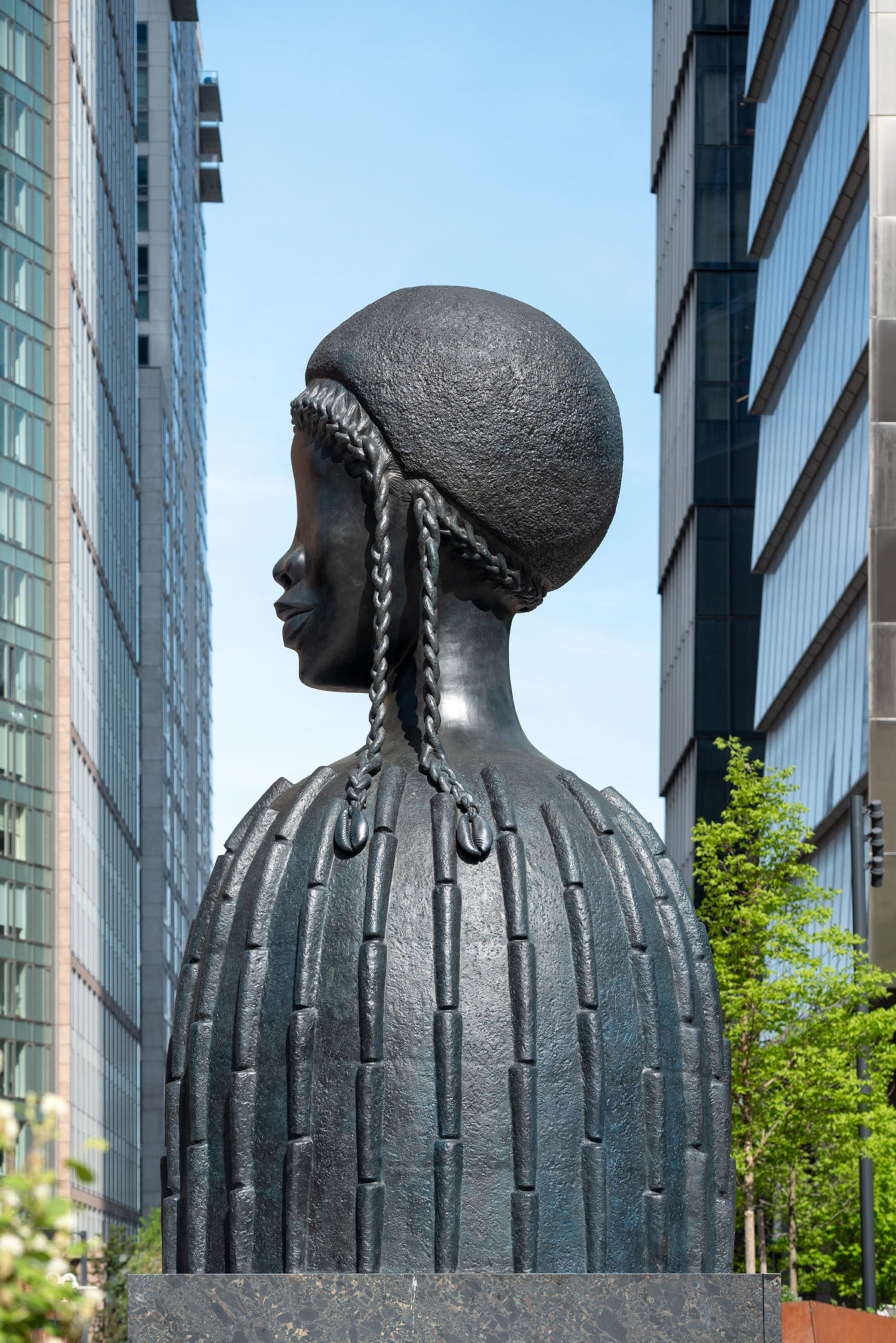 A large black sculpture of the head and torso of a figure. The figure has one singular braid wrapped around the edges of the head, and the rest of the hair styled in a small afro. The braids end with cowry shells. The sculpture is in between two rows of buildings.