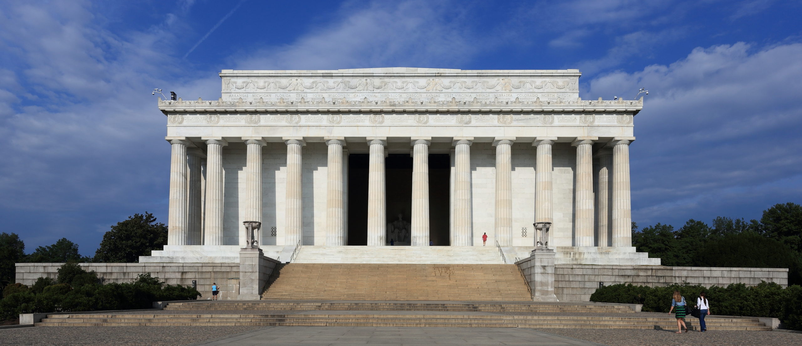 A wide image of the Lincoln Memorial (a horizontal building with Roman columns and descending stairs in the front), on a sunny day with a few bystanders walking around.