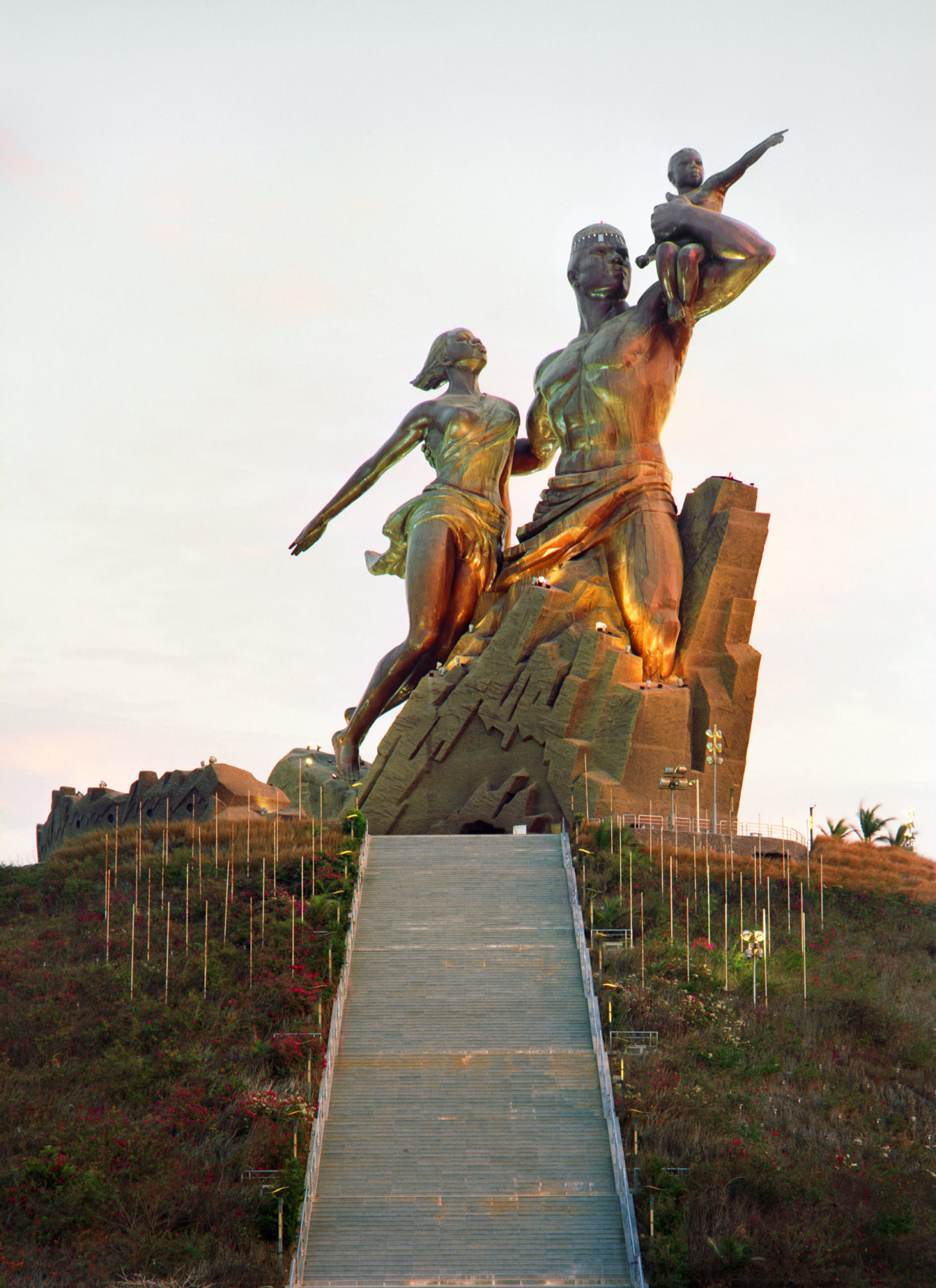 Copper statue atop of grassy hill with ascending stone steps; statue depicts larger-than-life figures of man with child upon his shoulders and a woman, their clothing blowing in wind, staring off into distance