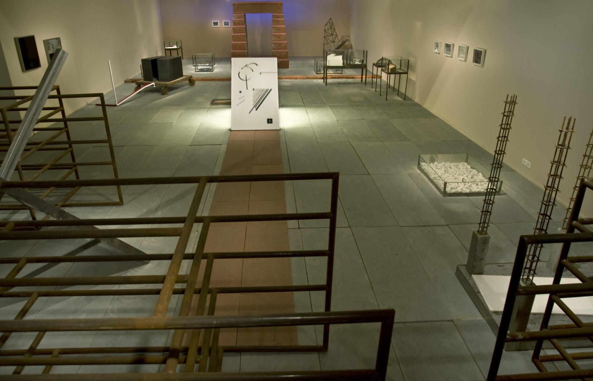 A large indoor gallery showing several artworks. In the foreground there are a series of fixtures which look diagrams, with perfectly measured rectangles atop each other, creating a three dimensional playground. In the center of the room is a brown paneled pathway which leads to a small white board covered in angled lines of different shades of gray and unfinished circles. Behind this is a large light brown altar which is surrounded by smaller pieces in glass tanks. There are several flat pieces framed on both sides of the walls.