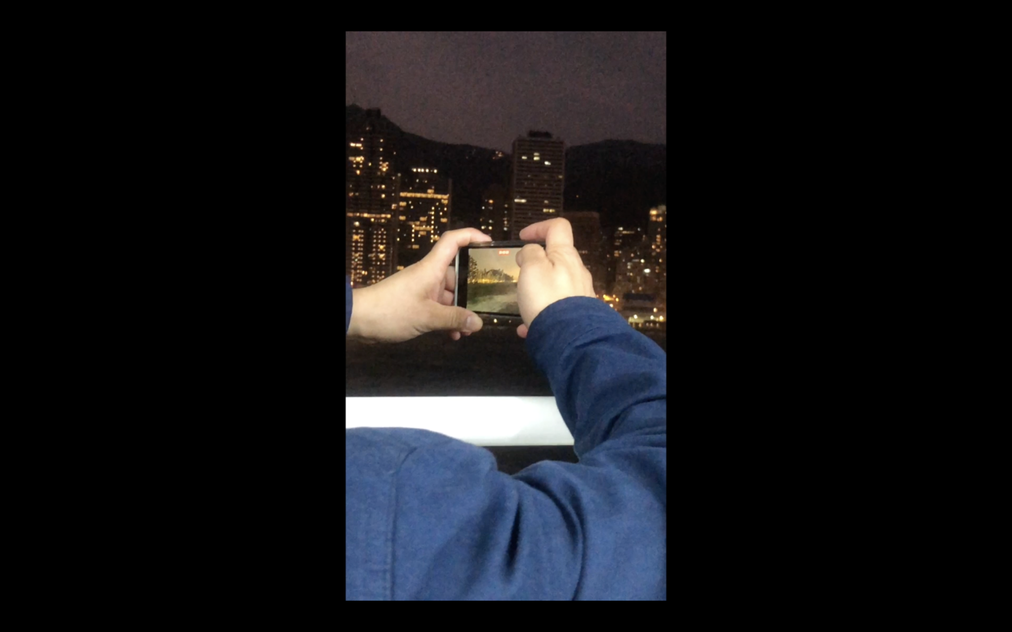 From the perspective of Sia's phone recording, we see two arms holding an iPhone over a white balcony and recording a protest scene that is offscreen to the left of frame. The skyscrapers of Hong Kong are seen in front of the person and their phone, and it appears to be dusk.
