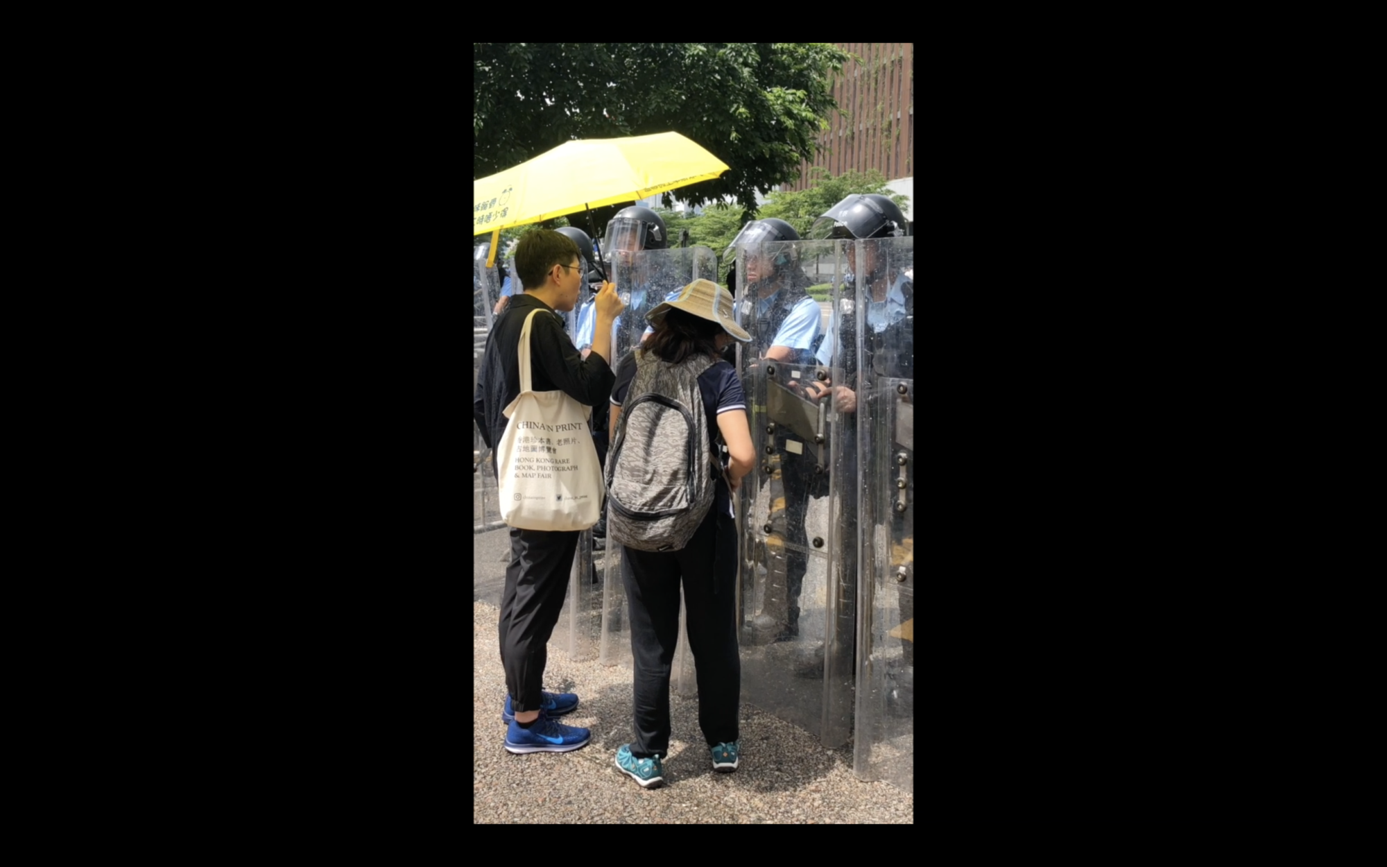 Two civilians speaking to a line of militant police officers who are holding transparent shields and blockading a building.