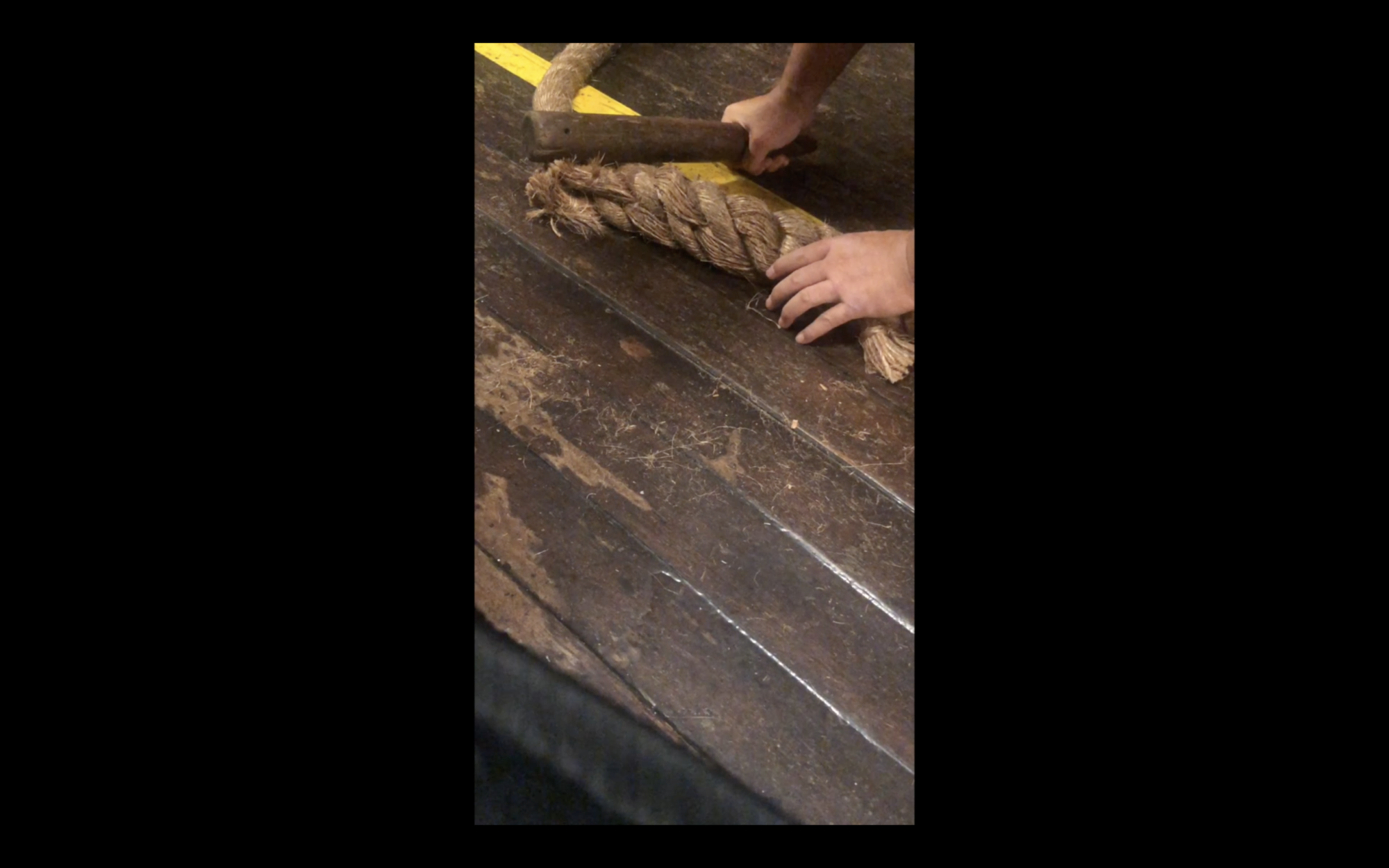 Video still of a pair of hands on a wooden floor handling a large piece of rope, and beating it with a wooden pallet.