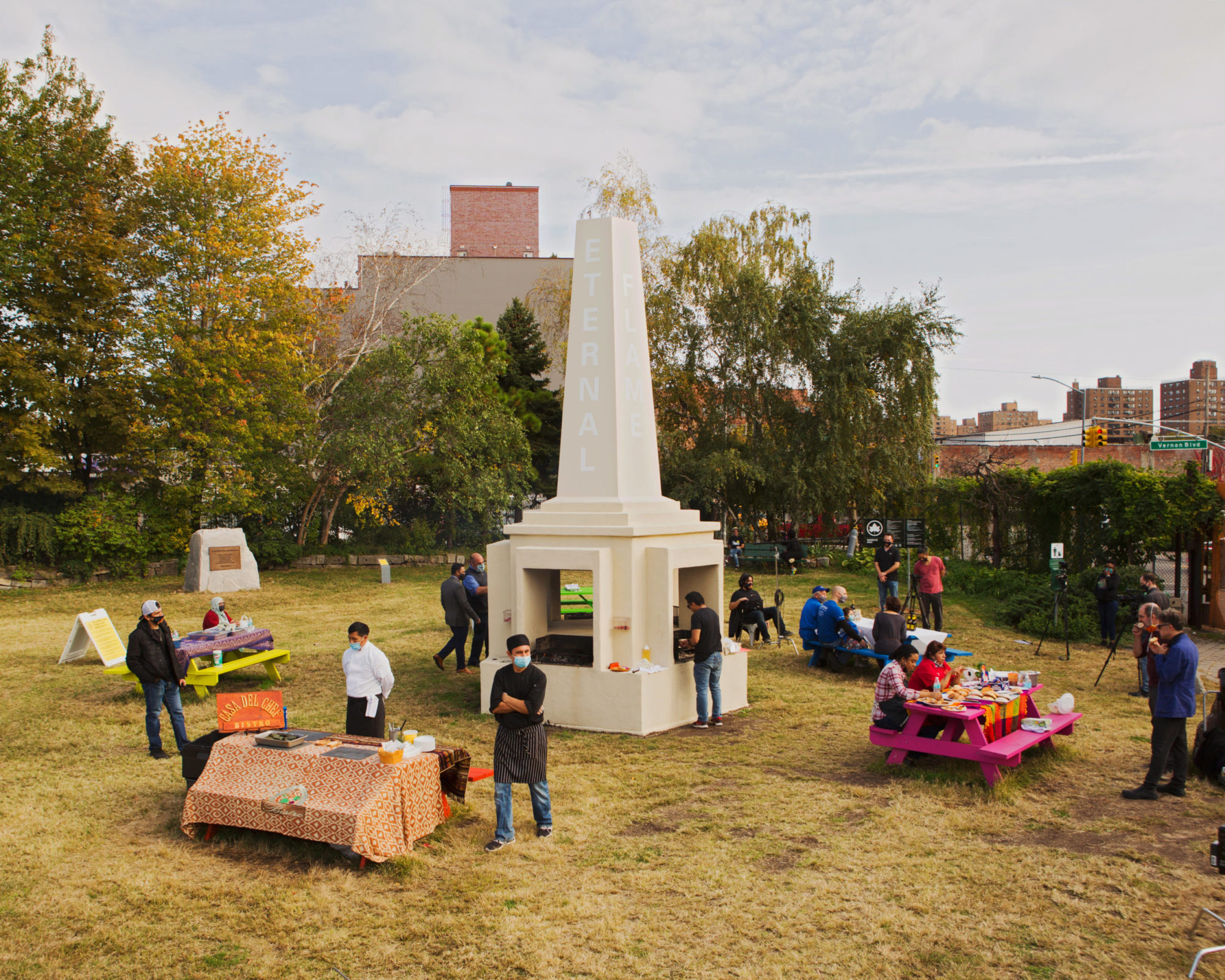 Community members milling around colorful picnic tables in grassy park, grilling on and around a faux monument with the words 