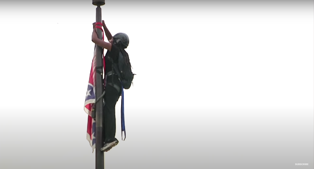 A person hanging atop a flagpole, dressed in all black with a helmet and backpack. They are reaching to remove a confederate flag.