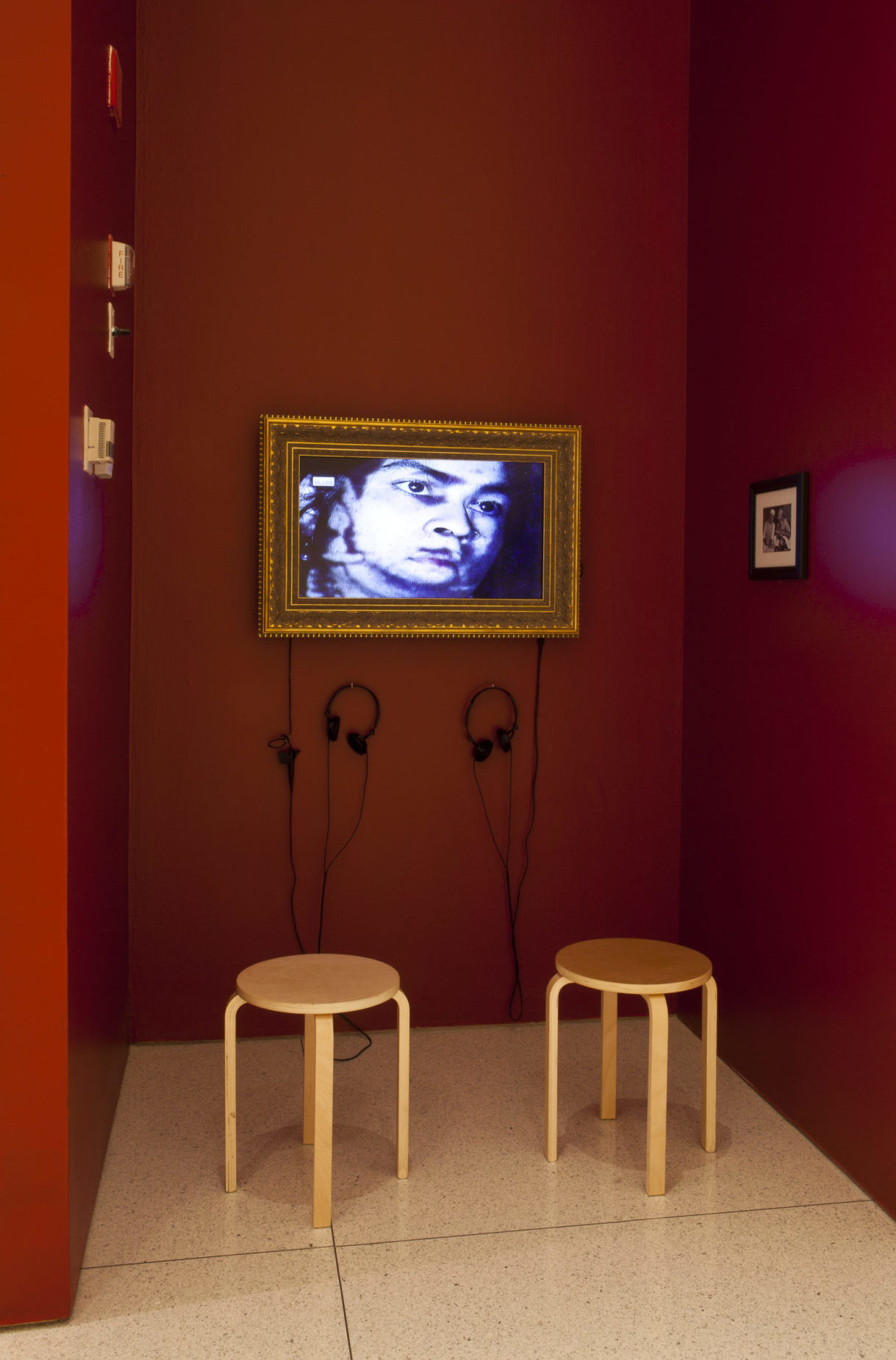 A small red room with a digital image hanging in the center of the front facing wall. The image appears to be a human face close up, and looking to the right and is framed in a gold border. Underneath the image is a pair of black headphones connected to it, and a pair of wooden stools for sitting. On both sides of the wall there are small pictures hanging up and framed.