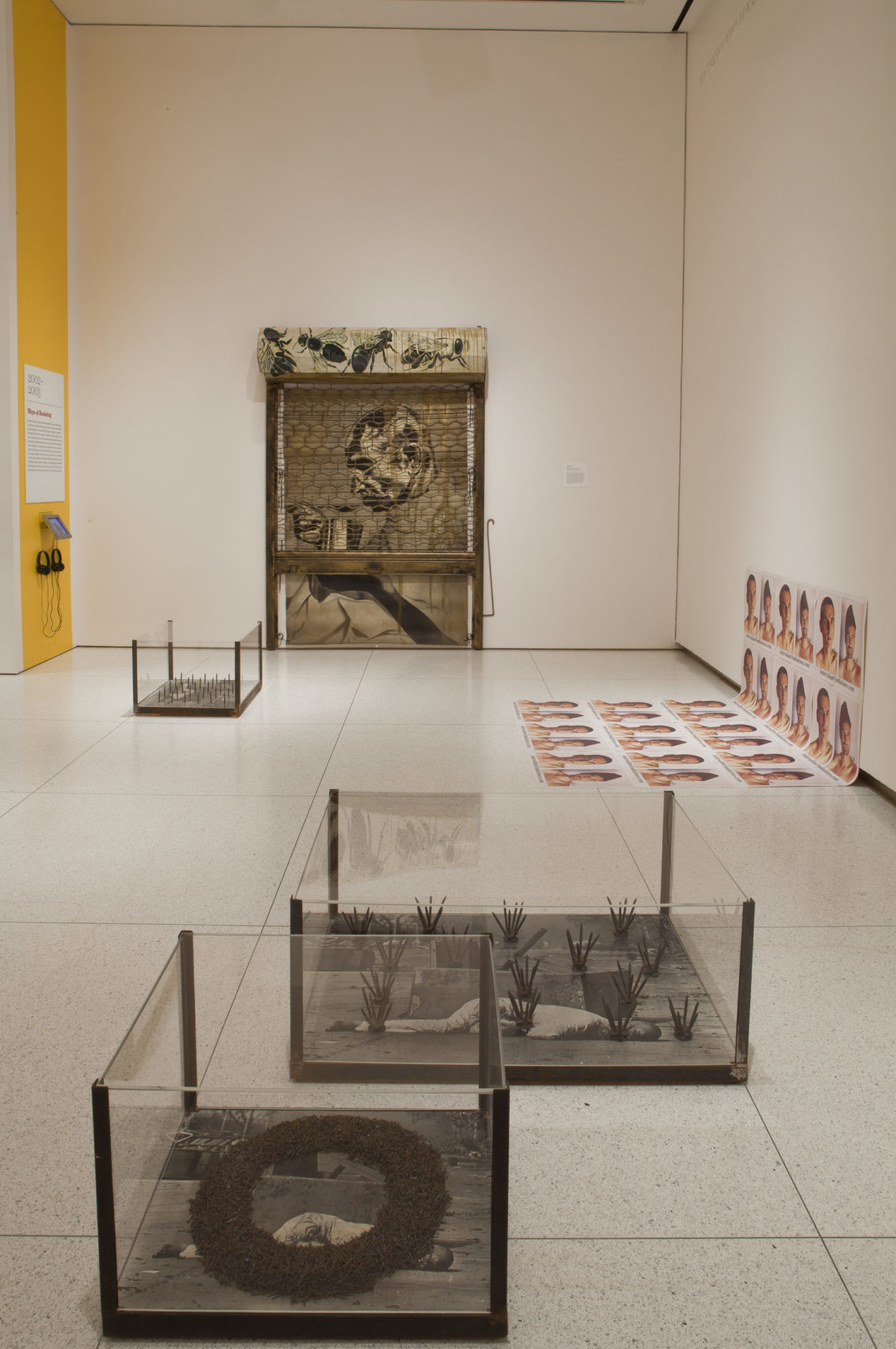 An open floor of a gallery room. On the front facing wall there is a back and white photgraph of an older man's left profile. In front of this picture is a sculpture of crosshatched vitrine of glass and iron. On the floor to the right of this, there's a single poster half slouching on the floor, which shows a series of headshots of someone in front of a white background. On the floor in front of the center picture, there are three glass tanks containing the same photograph of a man laying on the ground. Each copy is covered in arranged nails.