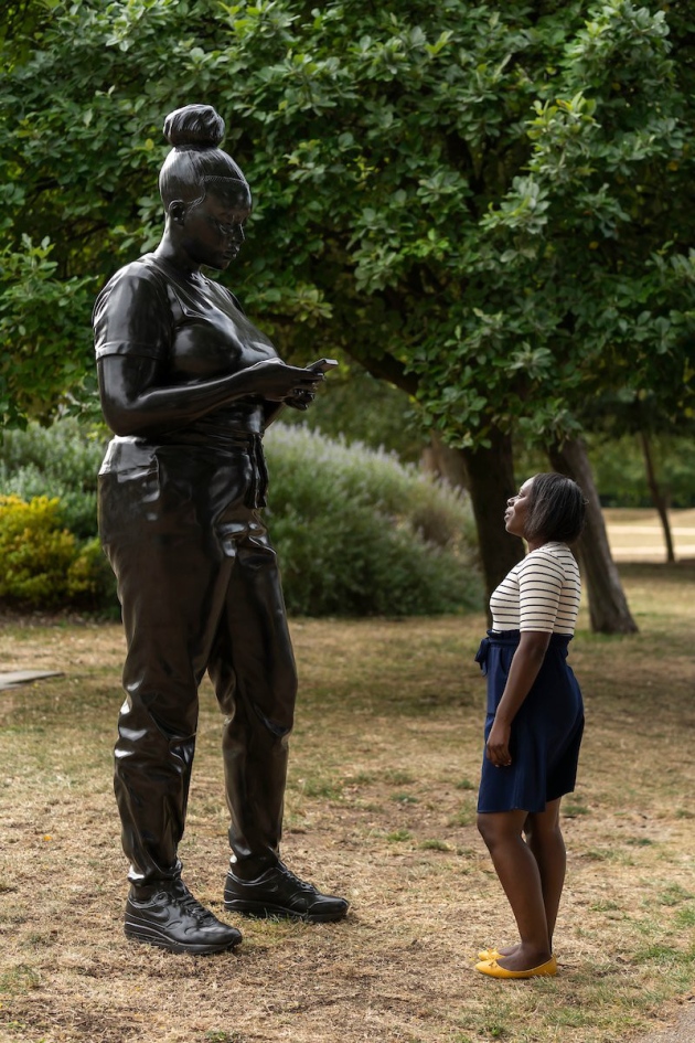 Young African American woman in jean skirt and striped shirt stands looking at dark bronze statue of African American woman in casual clothing and Nike sneakers, who is texting on her bronze cellphone