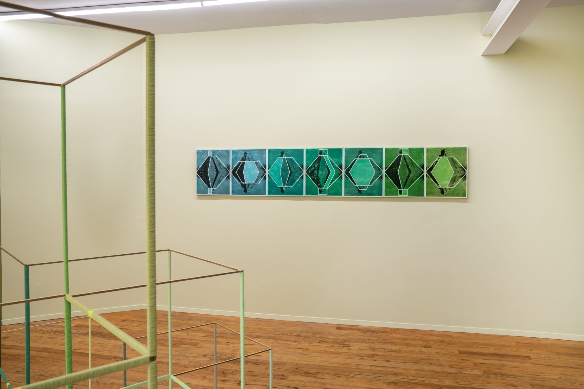 A series of seven wax panels with white lines creating geometric shapes over hues of blue and green. From left to right the blue fades into green, with the shapes changing in each panel meant to represent the movement of the sun on a water surface.