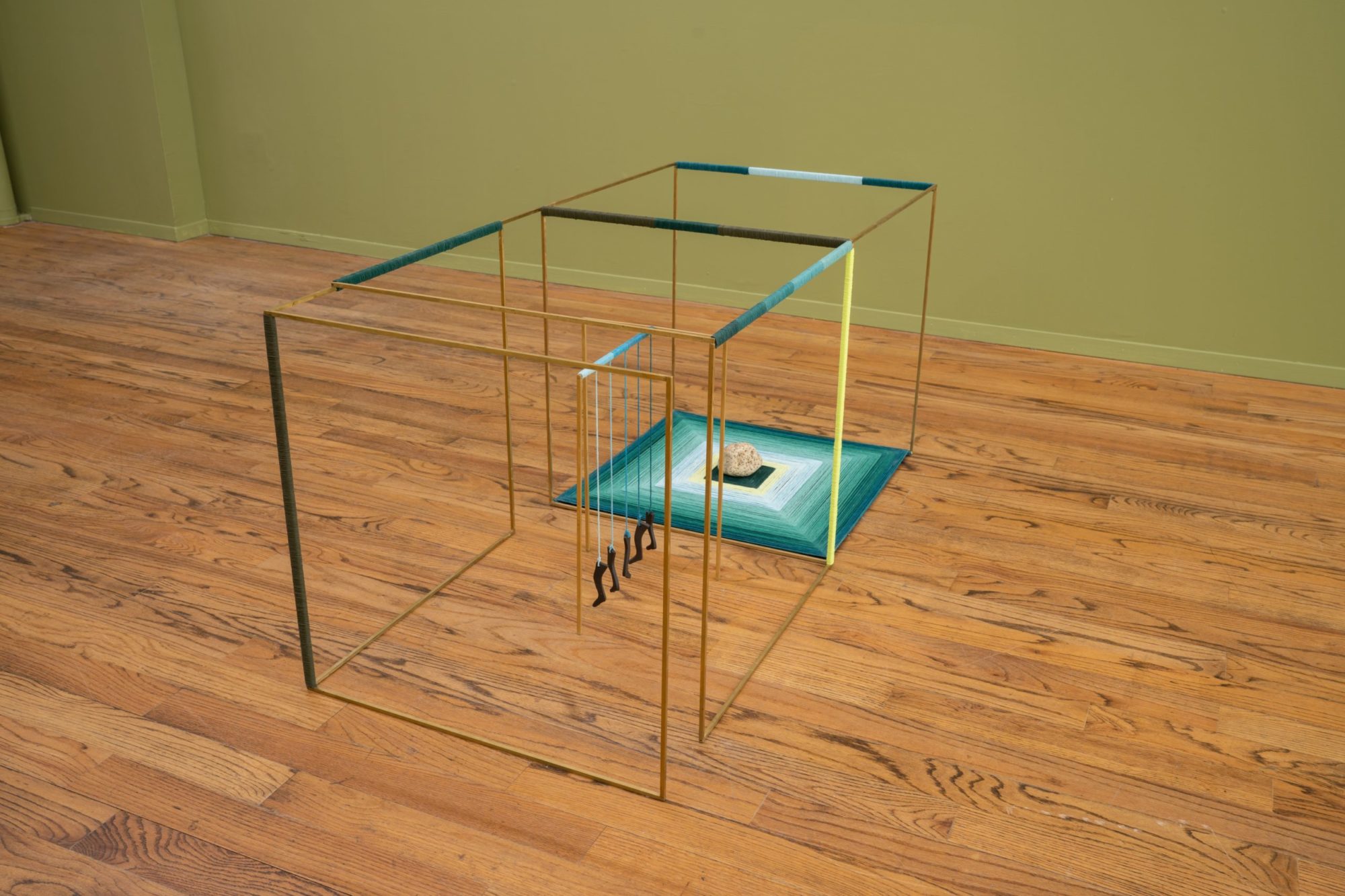 Single row of brass squares connected by blue and green threads creating two cubes. In the cube shape closest to the camera appears a row of river stones hanging by some blue threads. In the adjacent cube is a piece of rock sitting in the center of the cube's base. The base has squared layers of different threads colored blue, green, and yellow.