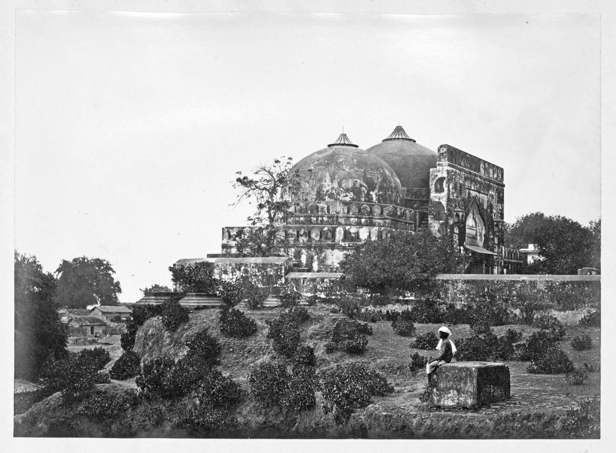 A black and white image of a large worn down building with two domes over the top. In front of the building there is sparse greenery, such as trees and bushes. Near the forefront sits a man with a white headscarf atop a stone.