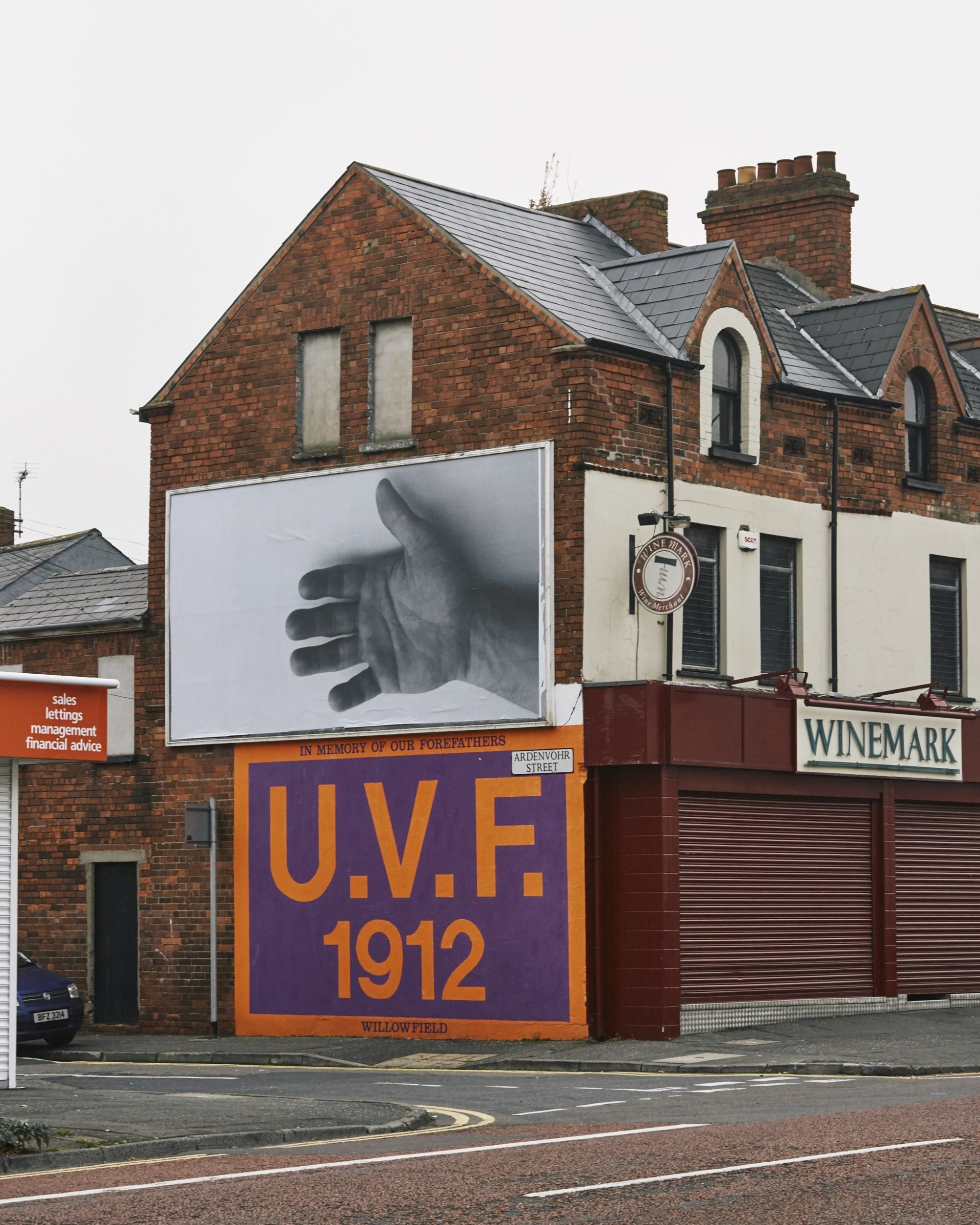 On the side of a tall brick building there is an a billboard featuring a black and white photograph of a hand, open and extending from the right side of frame. Underneath the display stands another signage which stated 