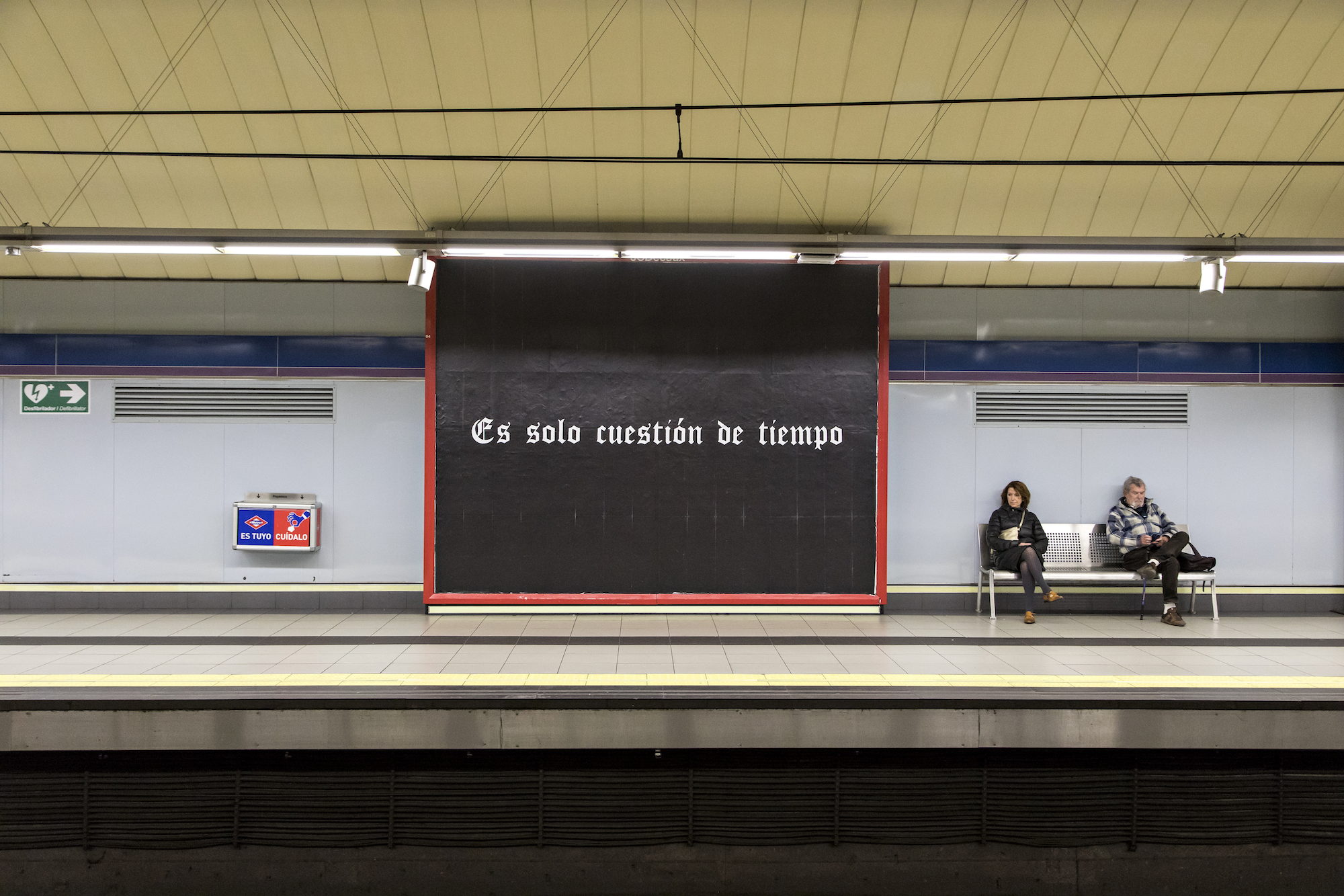 Inside a train station in which two bystanders are siting on opposite sides of a bench. To the left of the bench is a square wall displaying the phrase 