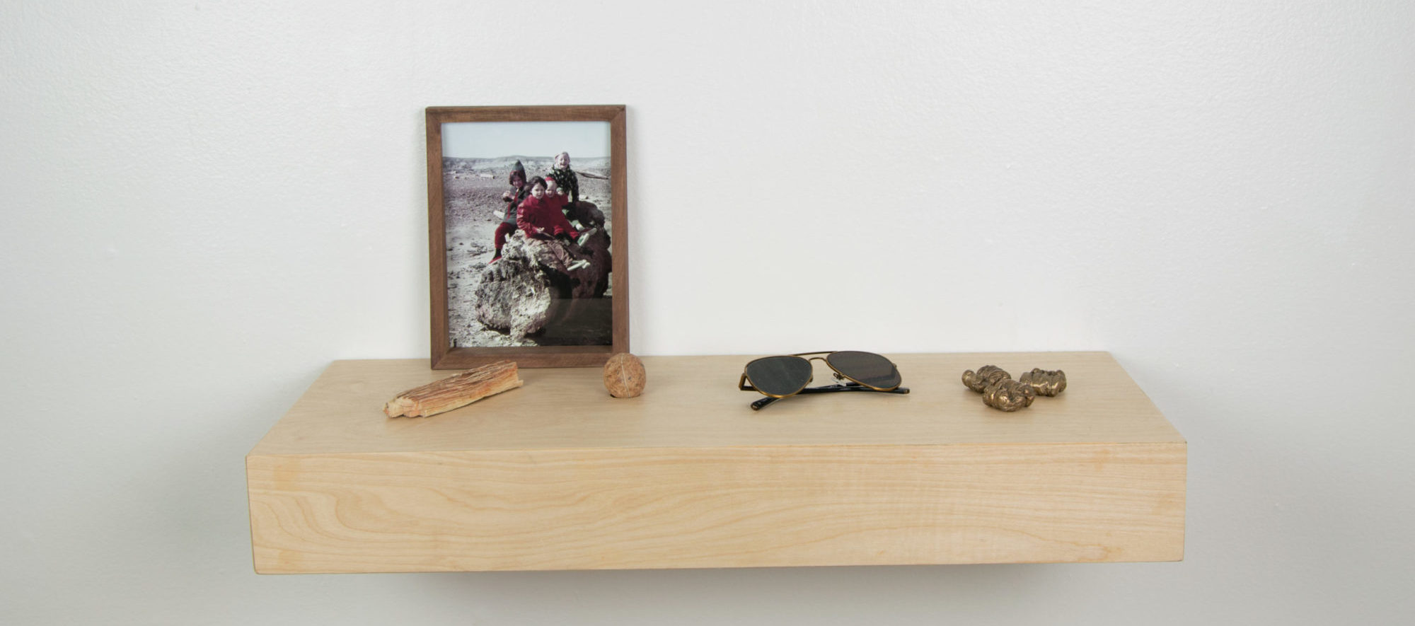 A wood fixture showcasing four objects (a collection of peanuts cast in bronze, a pair of black aviator sunglasses, one single brown rock, and a small light brown wood bark-like object) and a framed photo of four children sitting atop a large rock.