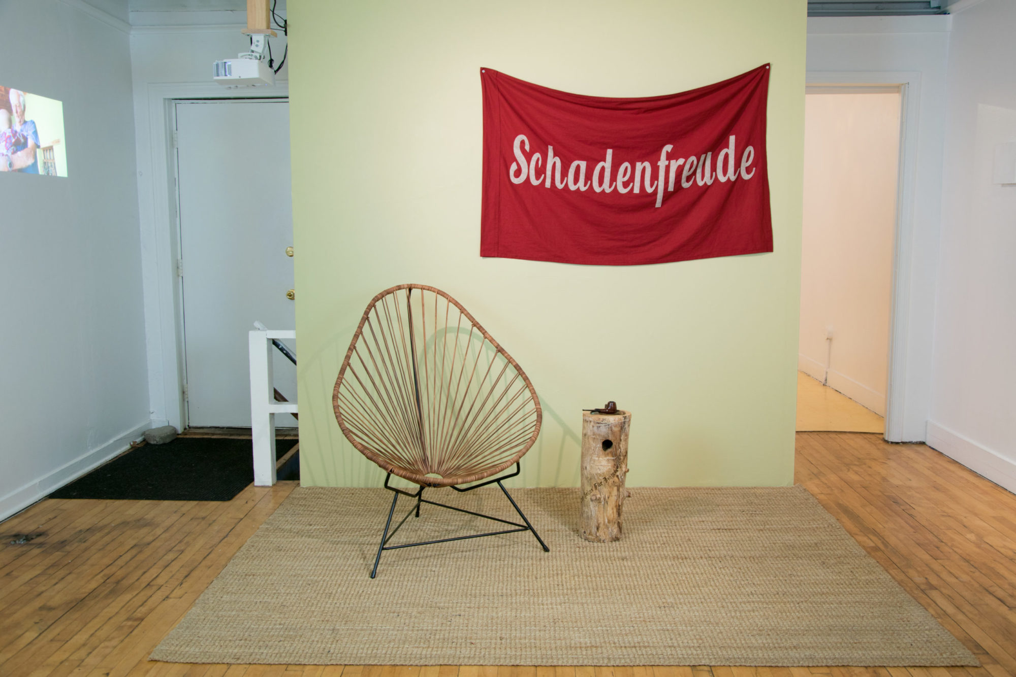 A sparse wick lounge chair is located beside a short stand made of light brown wood. The stand is underneath a red sheet hanging on the wall behind it, with the word 