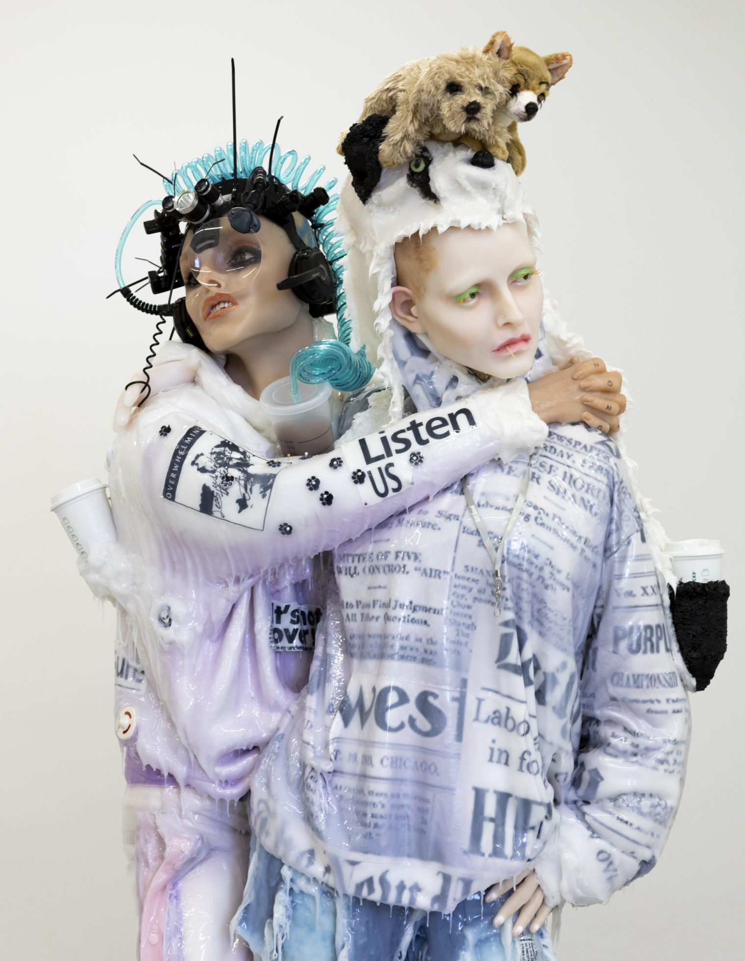 Two silicone figures dressed in white and pale-purple sweatshirts printed with text. Left-hand figure wears elaborate head-piece with plastic face-shield and curling plastic tubing in turquoise. Left-hand figure wraps arms around shoulders of right-hand figure, who stairs off, a white fur hood and two stuffed animals on their head. Both figures drip clear goop