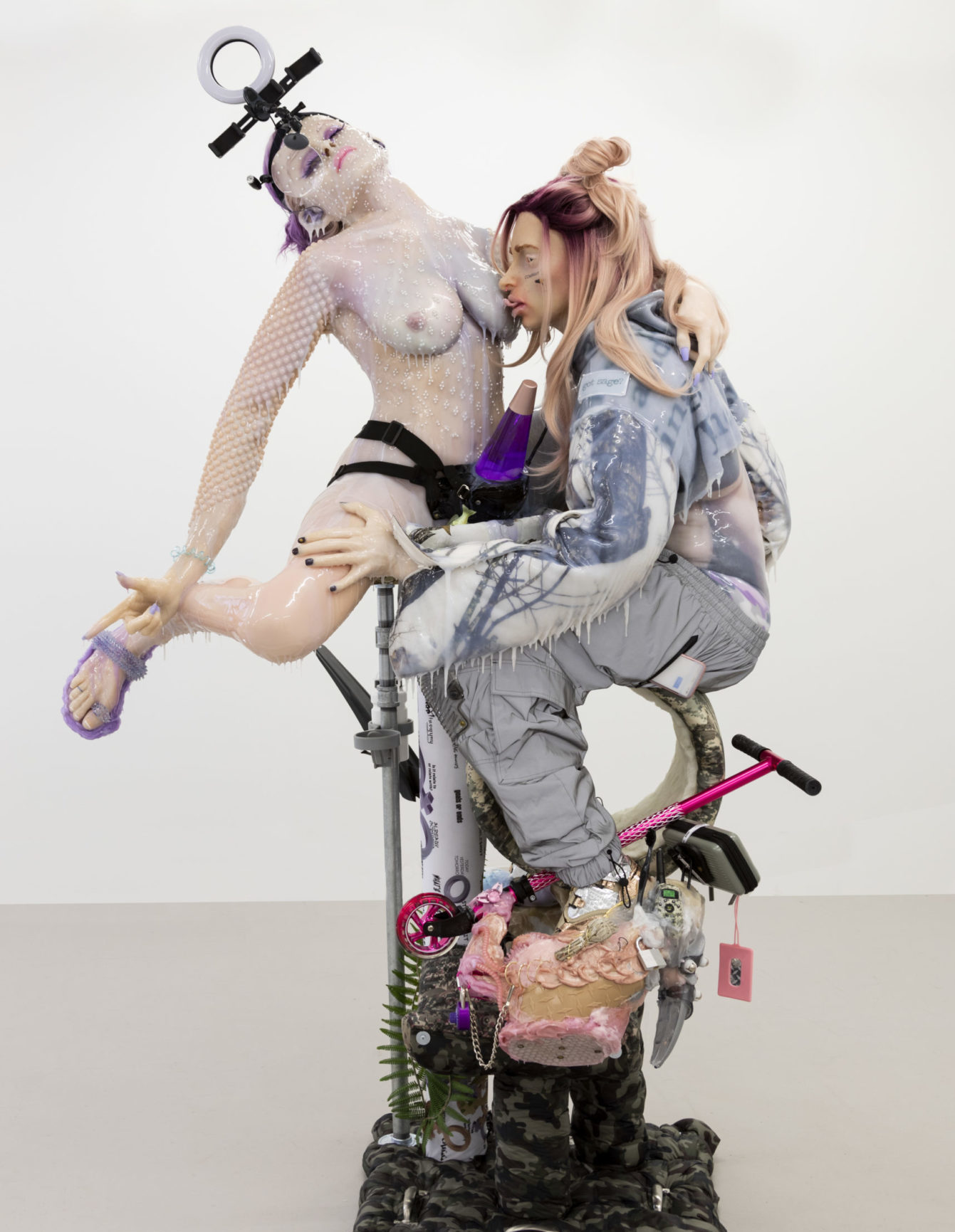 Two silicone figures on cloth pedestal. One figure, held up on metal struts and wearing black underwear and purple sandals, arches backwards as other figure sucks her nipple. Figure sucking her nipple wears tattered, acid-wash sweatsheart and loose gray pants. Both figures drip clear goop