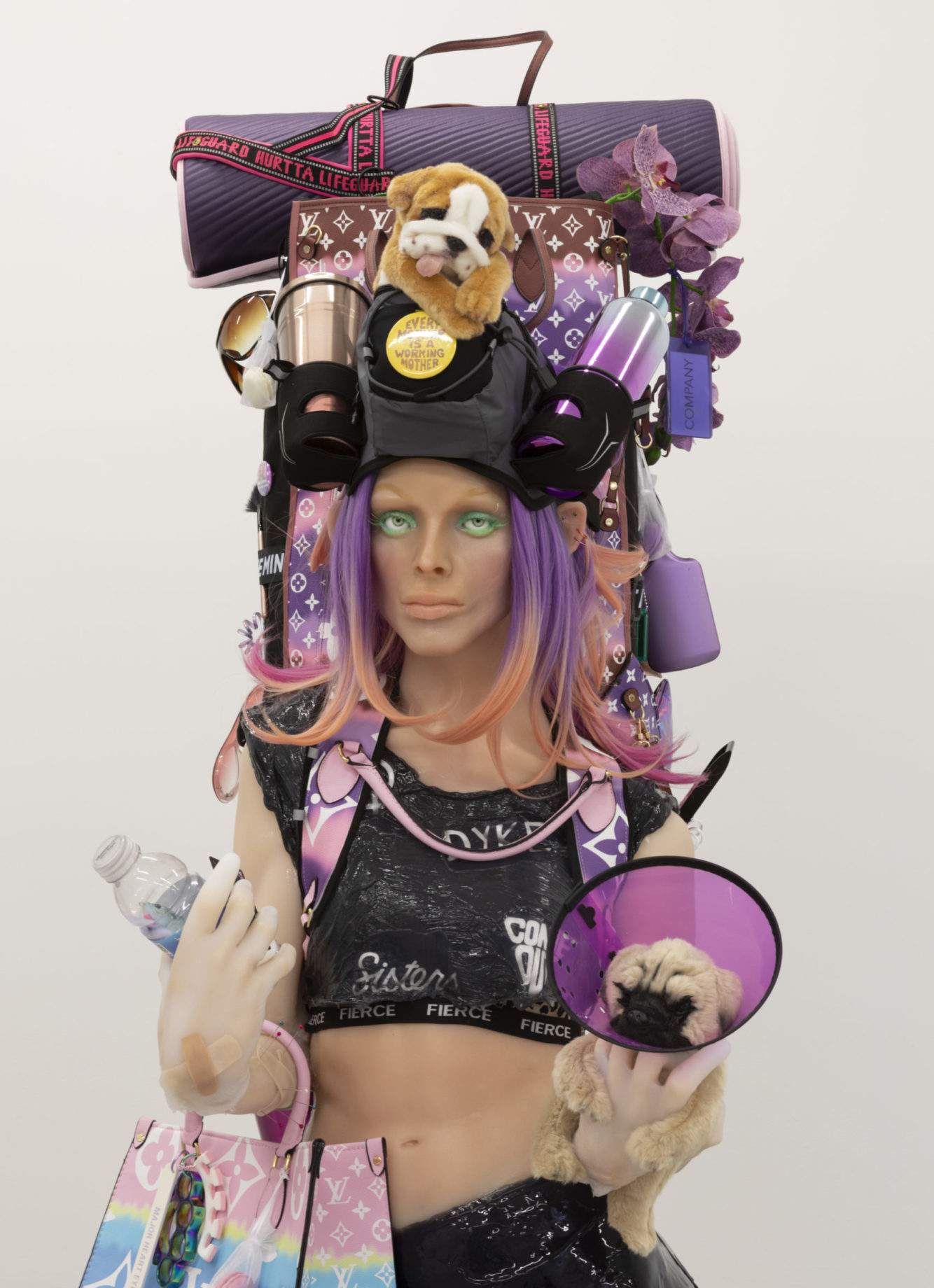 Spray-tanned silicon figure with bright green eye-shadow and purple hair stands iin black athleisure. On her shoulders is purple backpack decorated with designer brand logos and stuffed with waterbottles, portable coffee mugs, a yoga mat, a puppy, an orchid, and more. Mannequin holds another puppy in left hand and purse on right arm