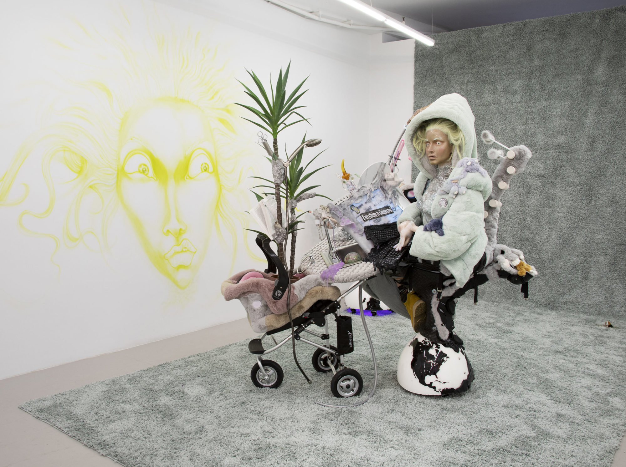 Silicone figure with wide eyes and bleach-blond hair sits with baby stroller against carpet backdrop. Figure wears faux fur jacket and is surrounded by items including a shower head, globe, and stuffed animals
