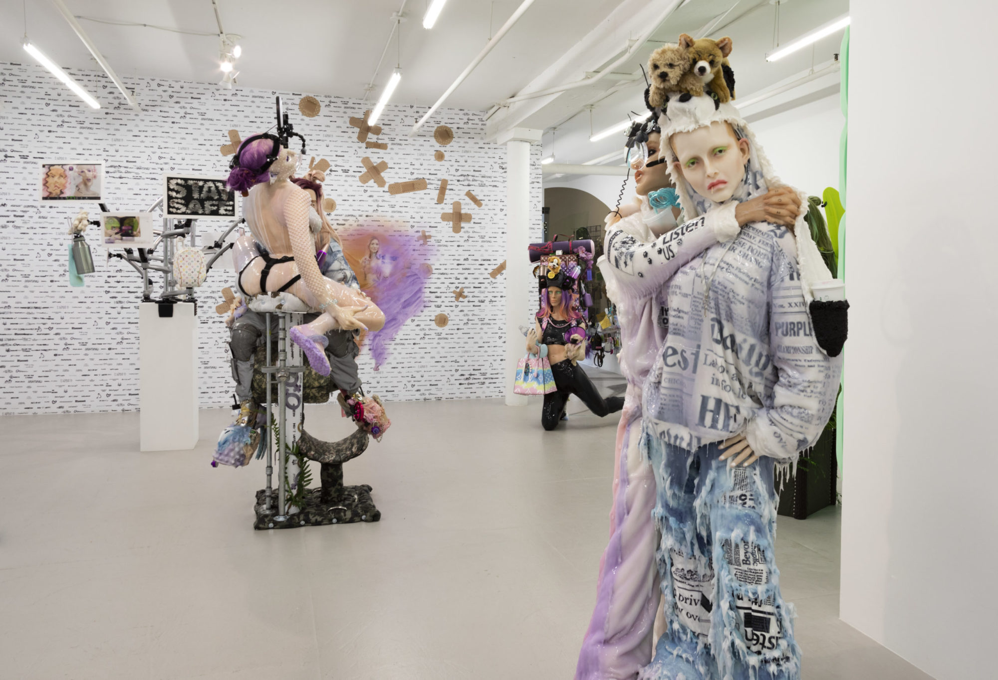 Three groups of silicone figures stand in various poses in gallery. On the left stand two figures in sexual pose; in back middle of frame kneels figure with huge purple backpack covered in accessories balanced on shoulders; to the right stand two figures, one's arms wrapped around the other, both dripping clear goo