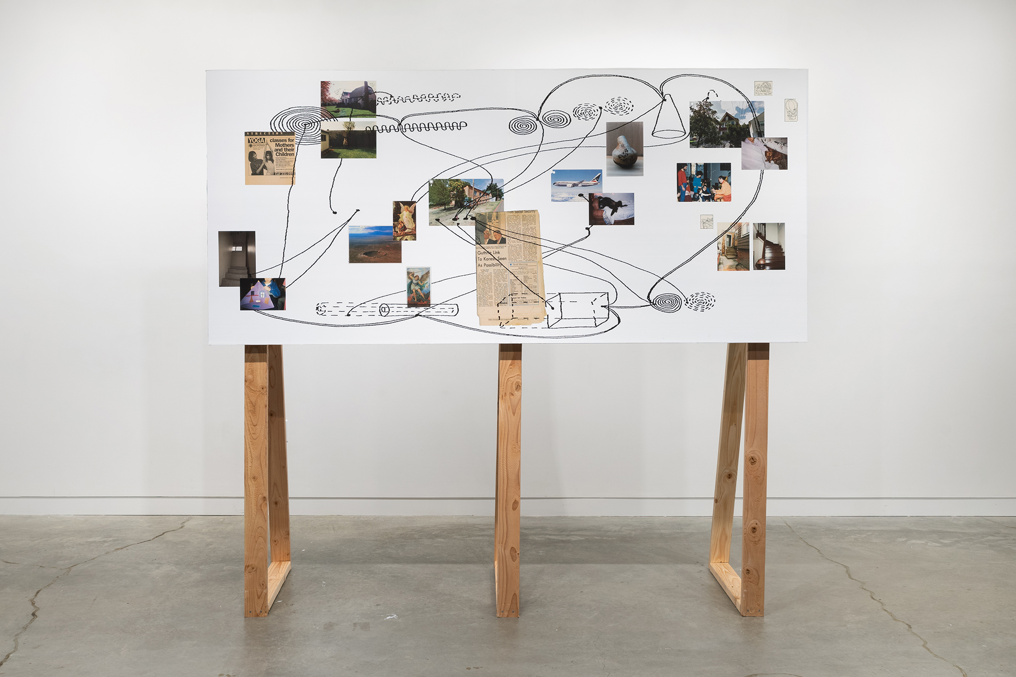 Long rectangular white board mounted on three wooden struts bearing photographs and newspaper clippings. Drawings and lines in black marker surround and connect the various clippings and photographs