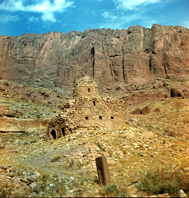 Photo of light brown brick shrine surrounded by rocks in Julfa cemetery, near collapse. The top is a dome structure, with several small windows on the outside, and an opening near the bottom left side.