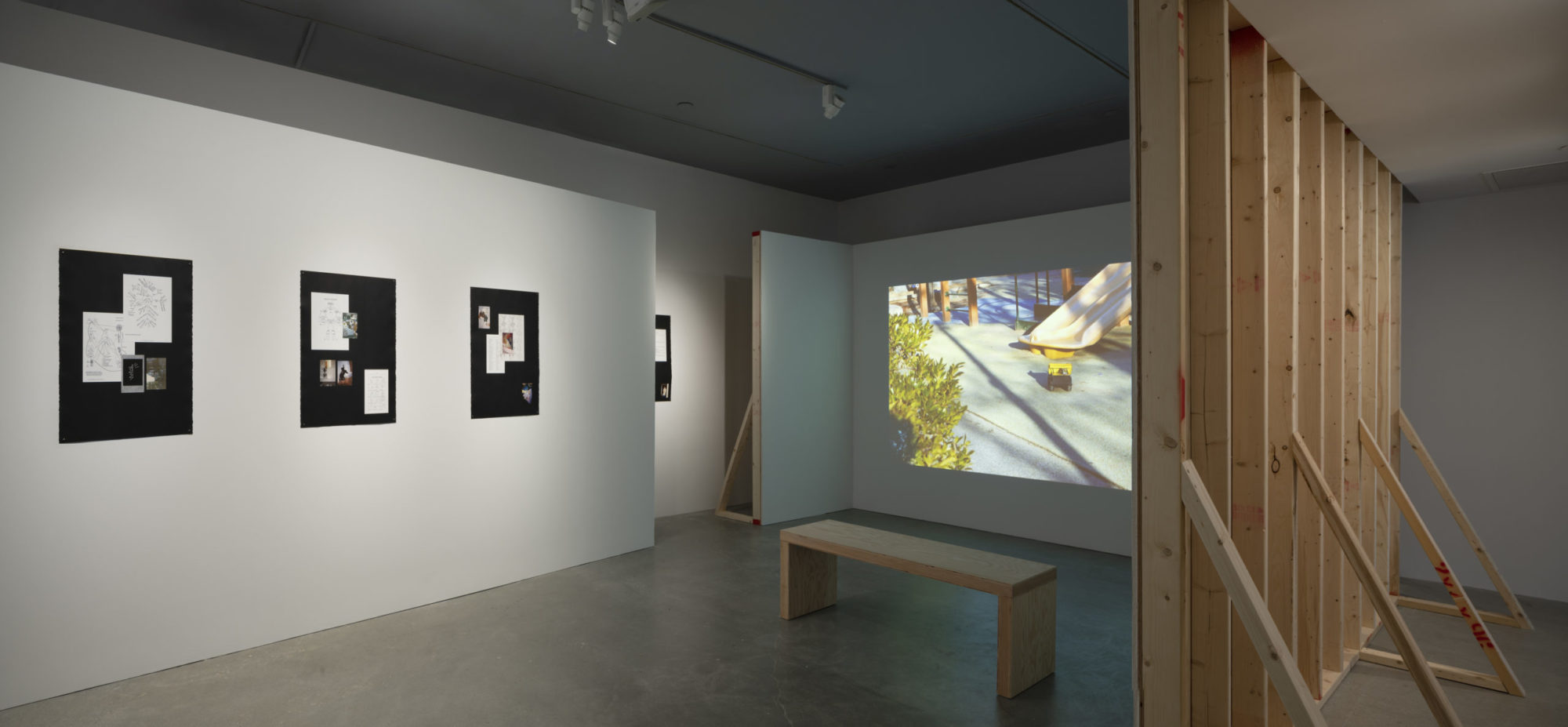 Gallery space showing vertical wooden construction beams, a wooden bench in front of a projected image of a children's playground slide, and a white gallery wall with works of art featuring pieces of ephemera upon black backgrounds