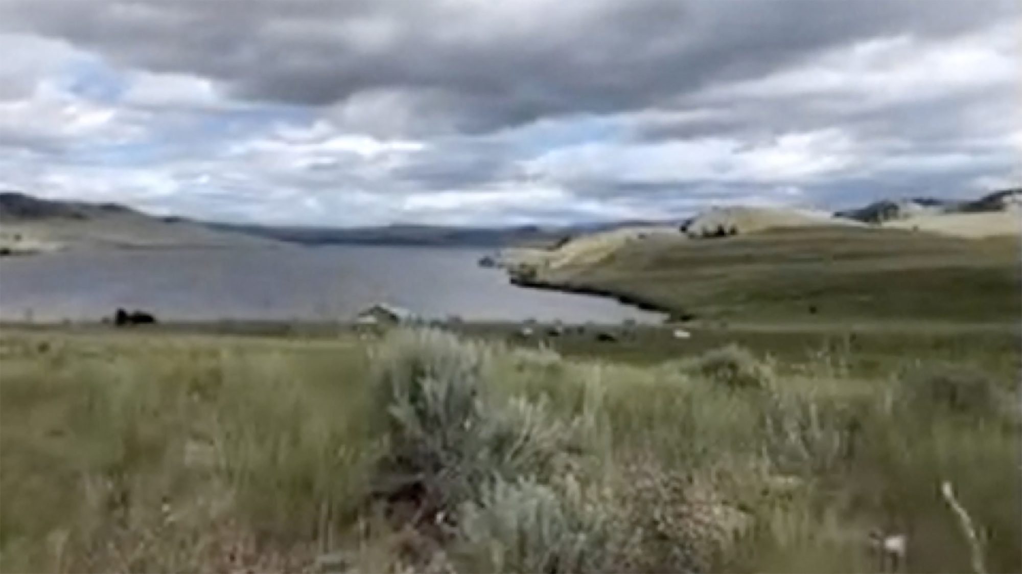 A landscape with a lake surrounded by a meadow under a cloudy sky — a patial view of the Syilx (Okanagan) Nation’s territory