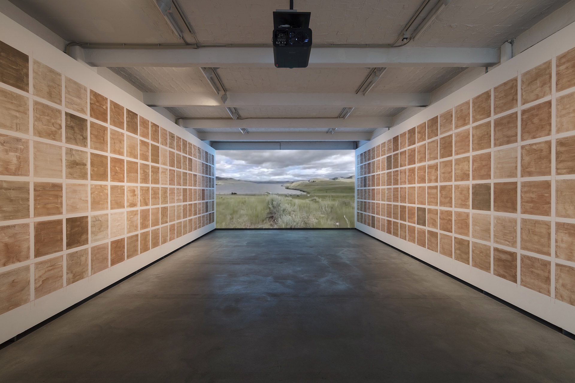 An image of a long gallery room with a projection on the far wall of a landscape with a lake surrounded by a meadow under a cloudy sky — a partial view of the Syilx (Okanagan) Nation’s territory; on the two side walls are grids of rectangles of different shades of brown