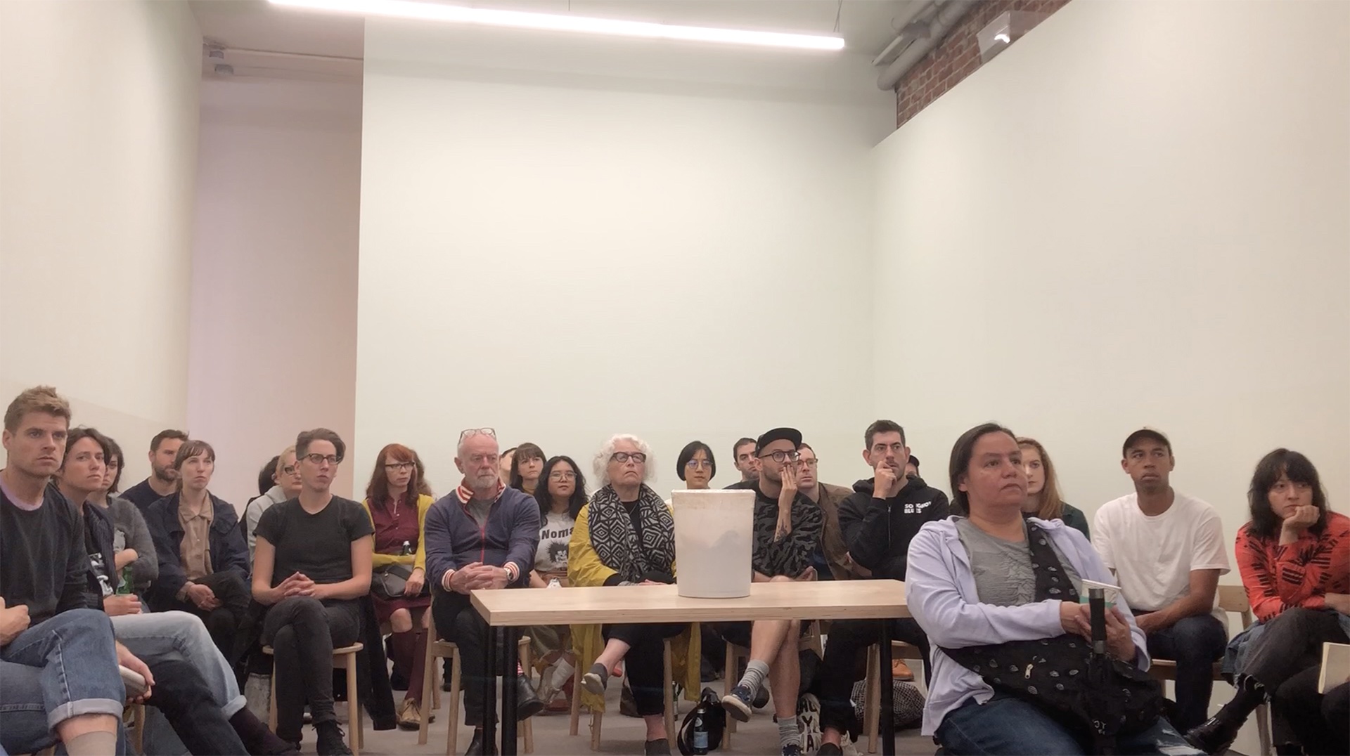 A white gallery room full of people sitting and staring past the camera; a wooden table stands in the middle of the audience