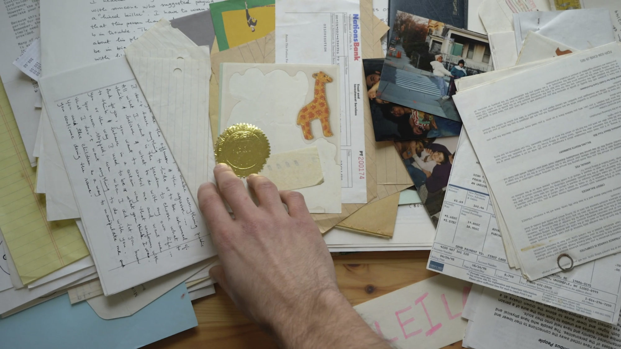 Image of a hand touching a pile of ephemera including stickers, photographs, old lettrs, certificates, and offiicial-looking documents.