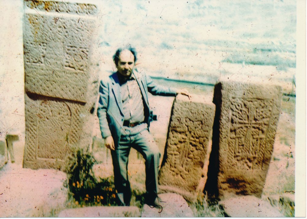Old photograph of Argam Ayvazyan standing next to three recovered Armenian Khachkar monuments all standing on ground.