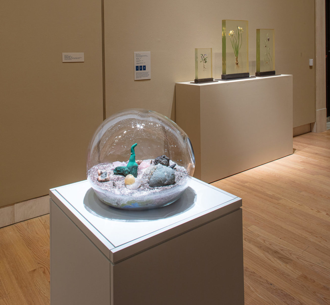 Installation view of podium holding a glass bubble encasing a small amount of sand and rocks. It includes small statues inside it as well.