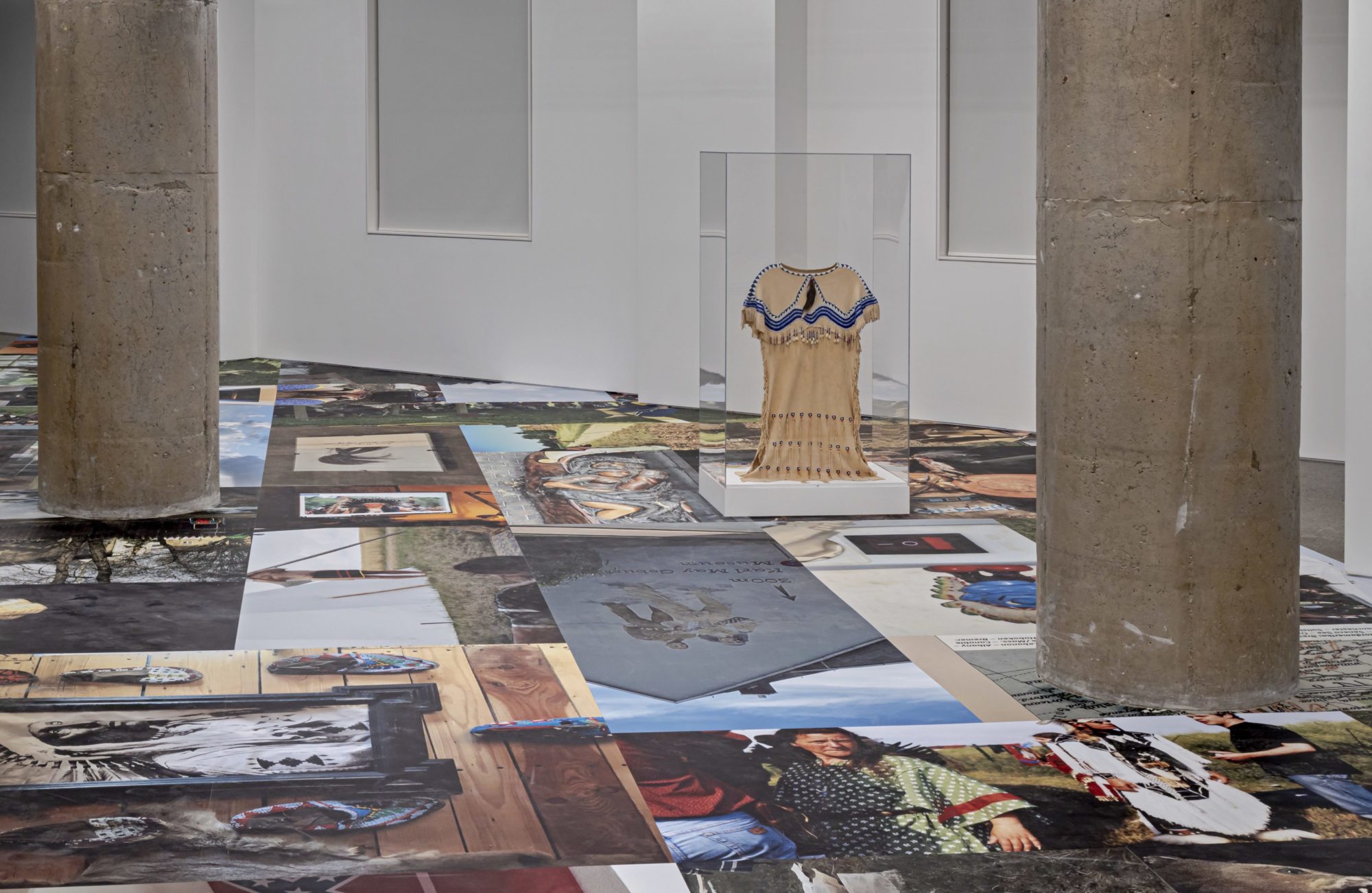 An image of a gallery room with a glass case containing a traditional Syilx (Okanagan) Nation dress; large photographs with scenes of Syilx people cover the gallery floor