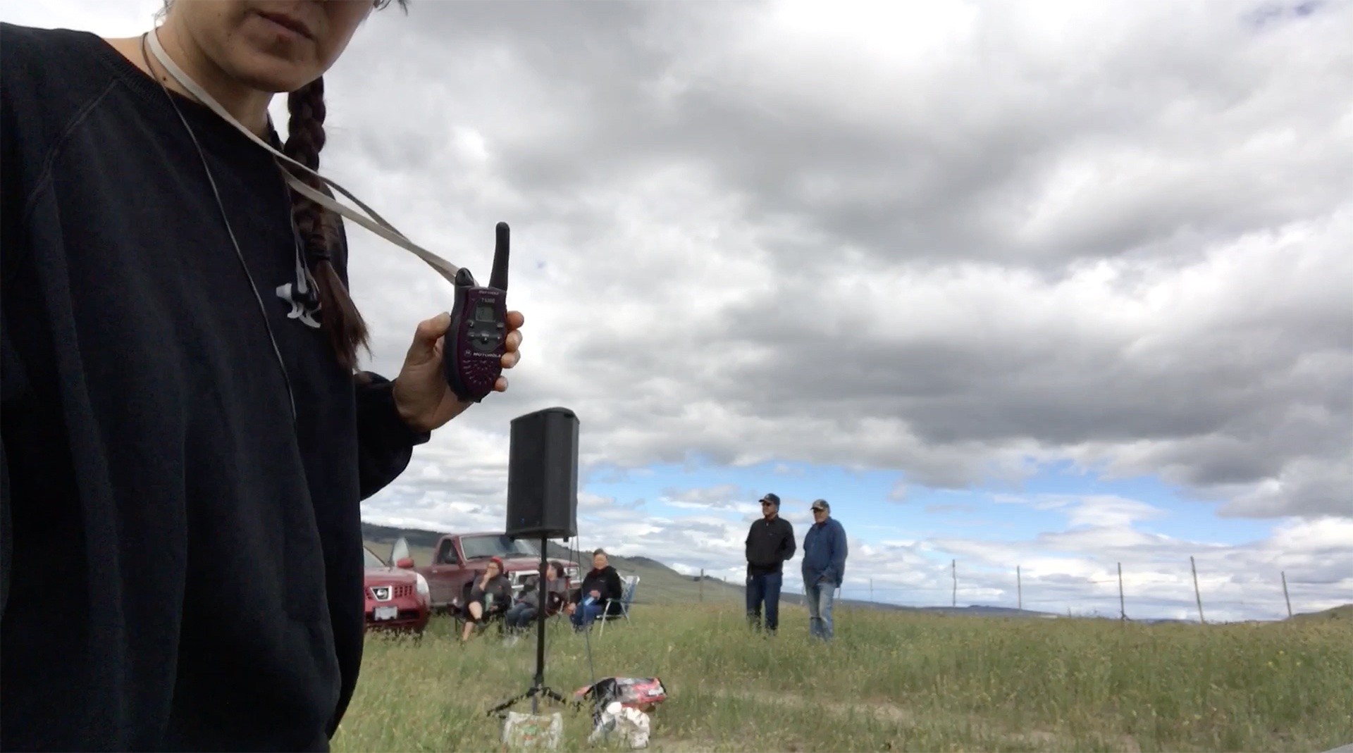 An image of artist Kista Belle Stewart holding a walkie talkie in a field on the Syilx (Okanagan) Nation’s territory; five people stand and sit on the grass in the distance, beside two red cars; a PA speaker stands in the middle of the field