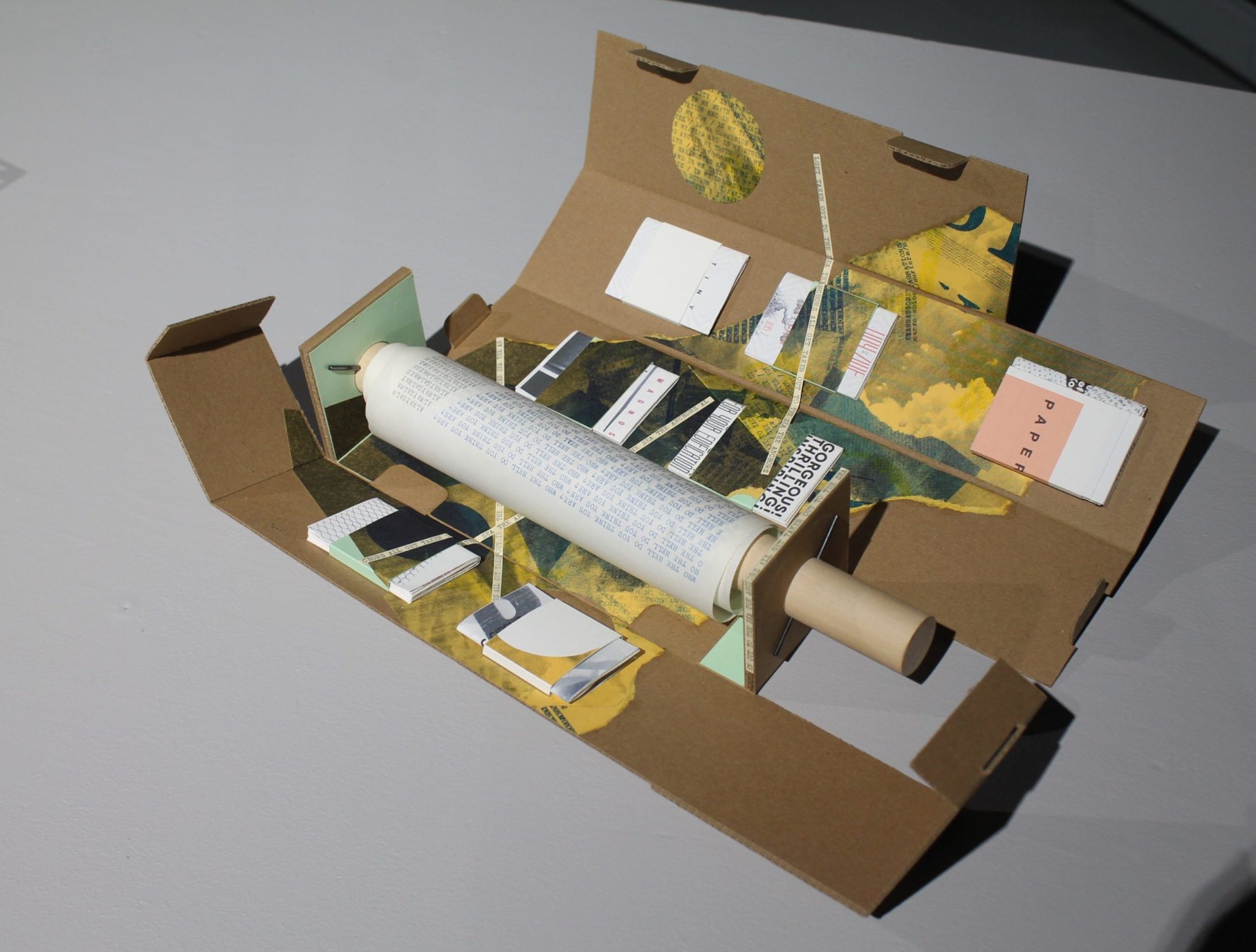A closer view of Thad Higa's Portable Library #8. The work s mixed media and is comprised mostly of paper products and collage. Although mainly neutral in color, there is some yellow throughout.