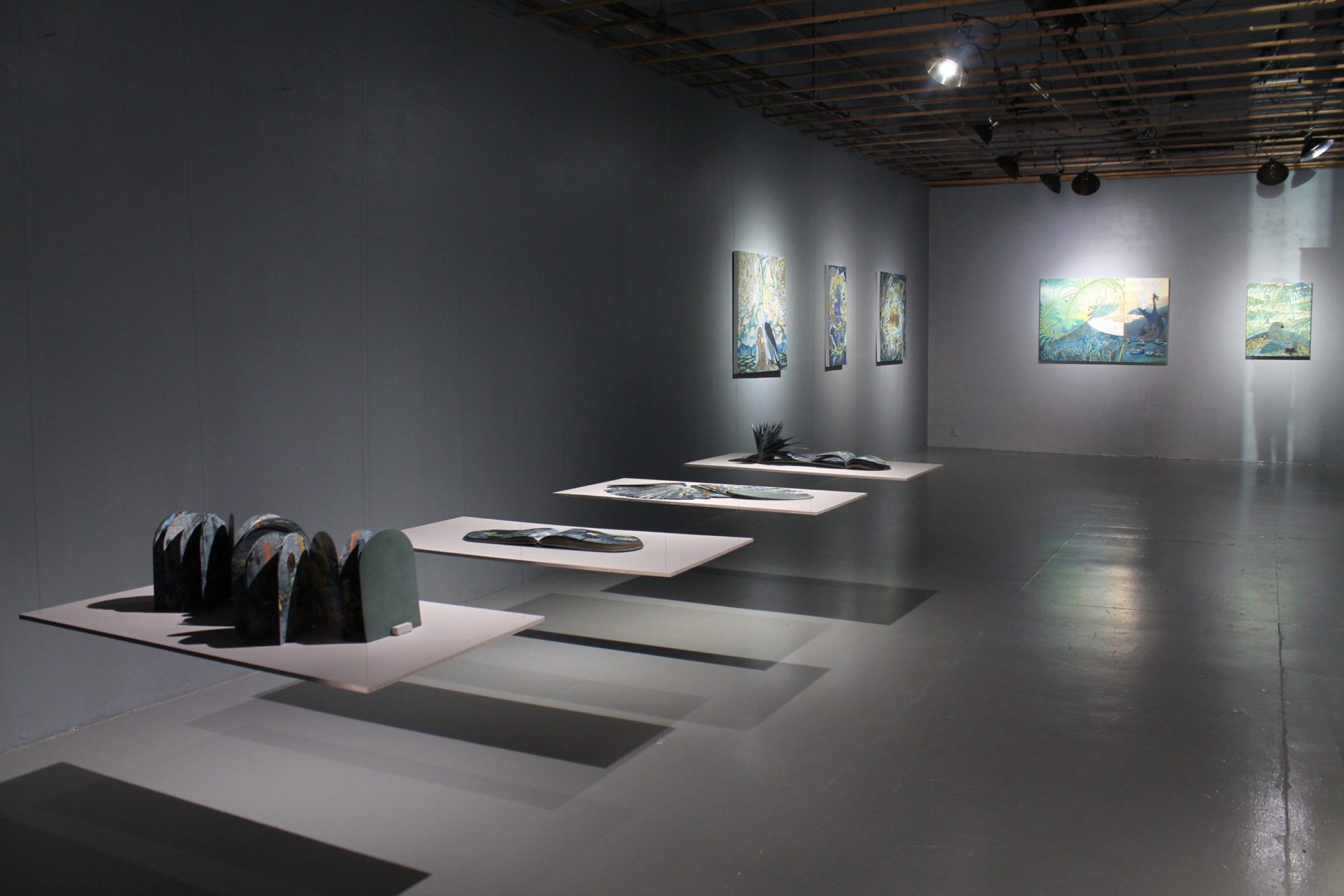 A wider gallery view focusing on Tammy Nguyen's artist books. The four artist books are on floating tables suspended from the ceiling along the left wall in view. Each is on its own table. Farther down the left wall are three paintings on the same wall and two paintings on the back wall. The walls, floor, and tables are all grey.