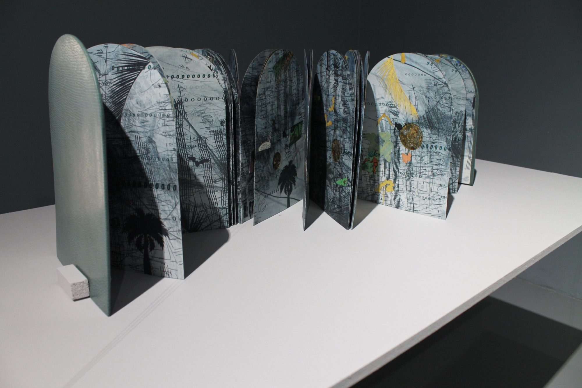 A closer view of Tammy Nguyen's artist book Four Ways Through A Cave #1. The book is arched in shape and is grey blue in color. The pages feature layered images and words and fold in on one another in an accordion fashion. Some pages feature additional mixed media.