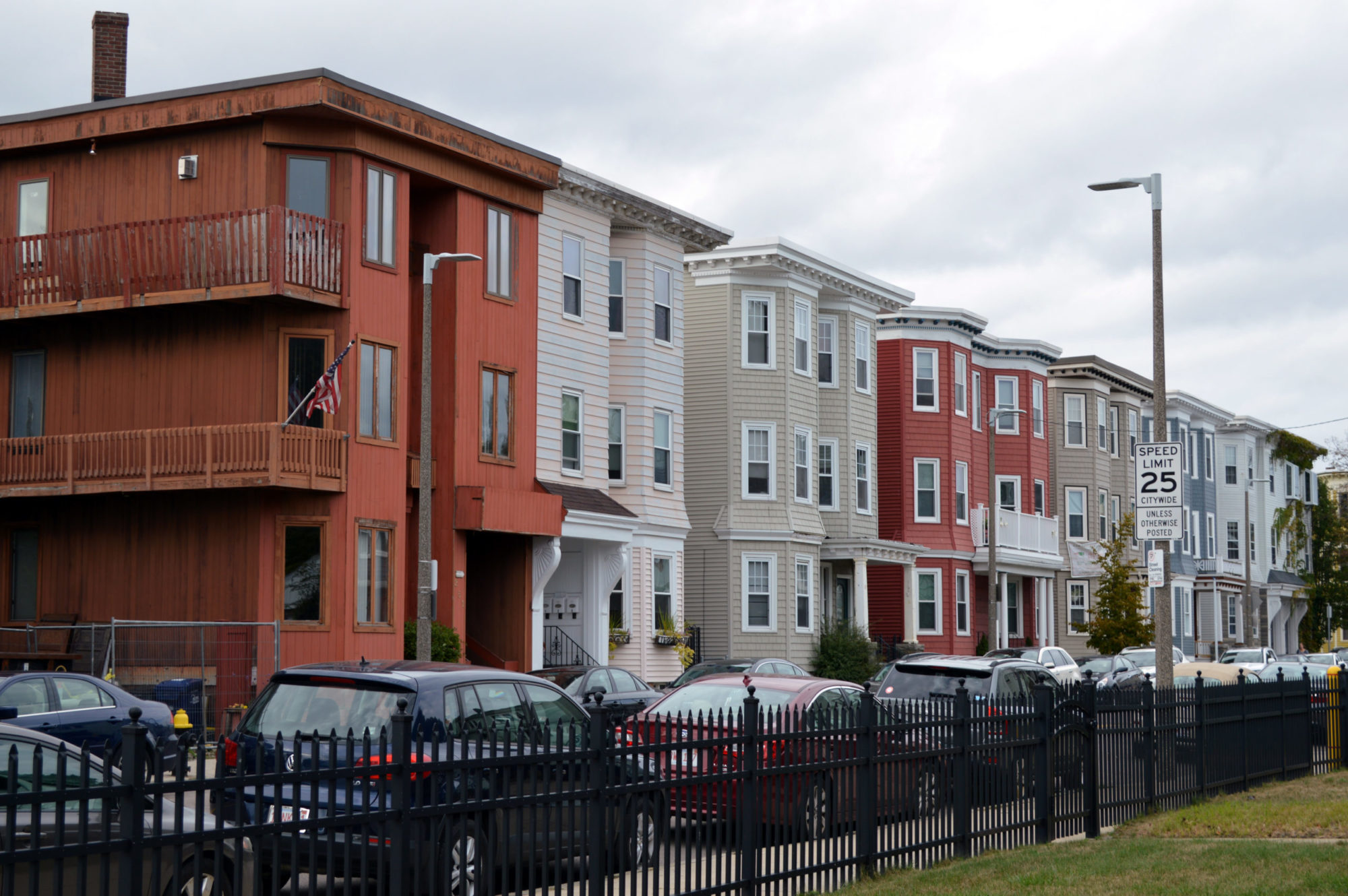 A street view shot of multiple three story homes attached to one another. Each building has a bay window and is a different color than the ones on either side. There aer cars parked along either side on the street.