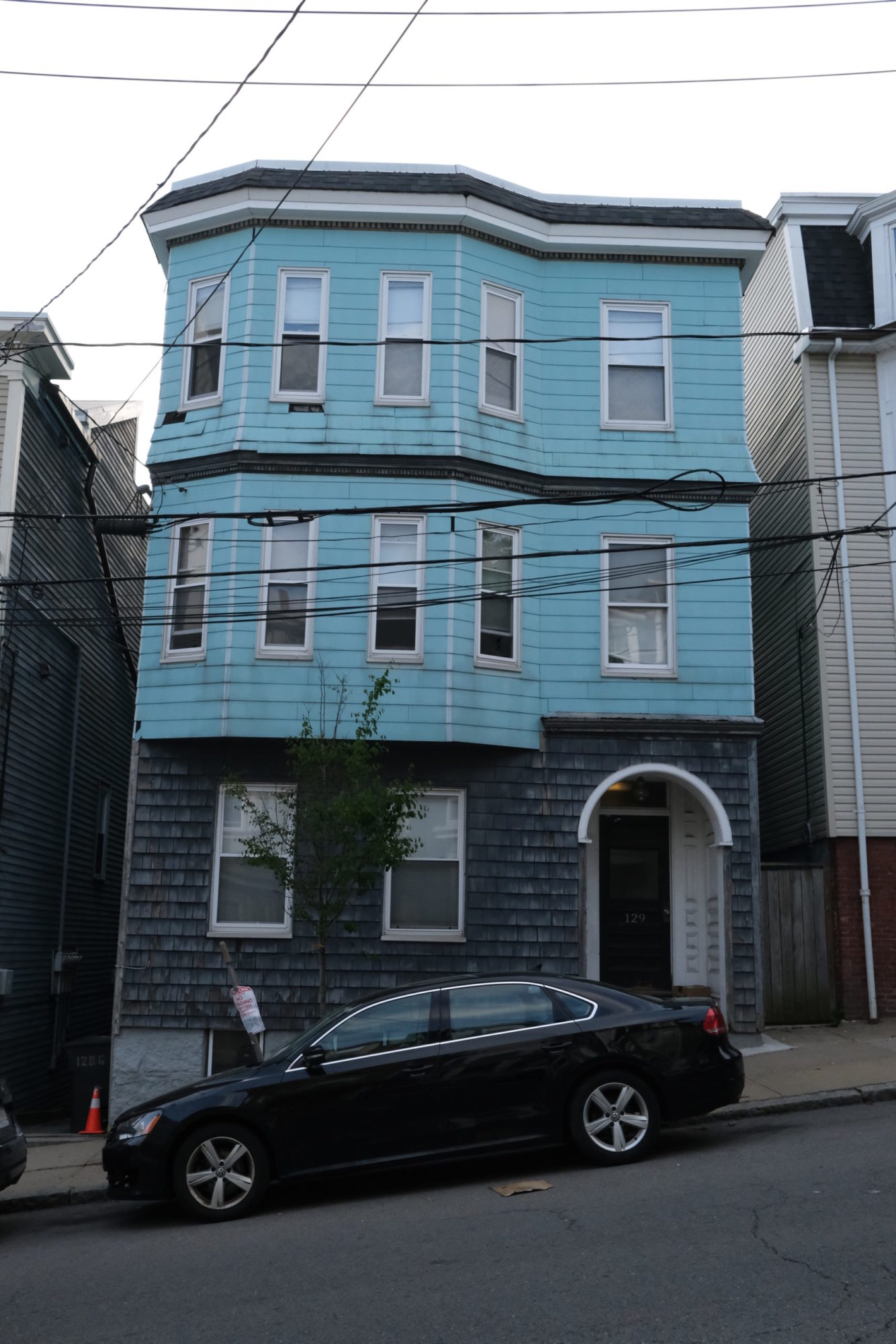 A straight on street view shot of a three story home. The bottom floor is dark grey. The top two floors are light blue. The front door is under an arched awning on the front right of the building. There are bay windows on the front left of the second and third stories. A car is parked directly out front. A street sign is bent. Telephone wires cross in view.