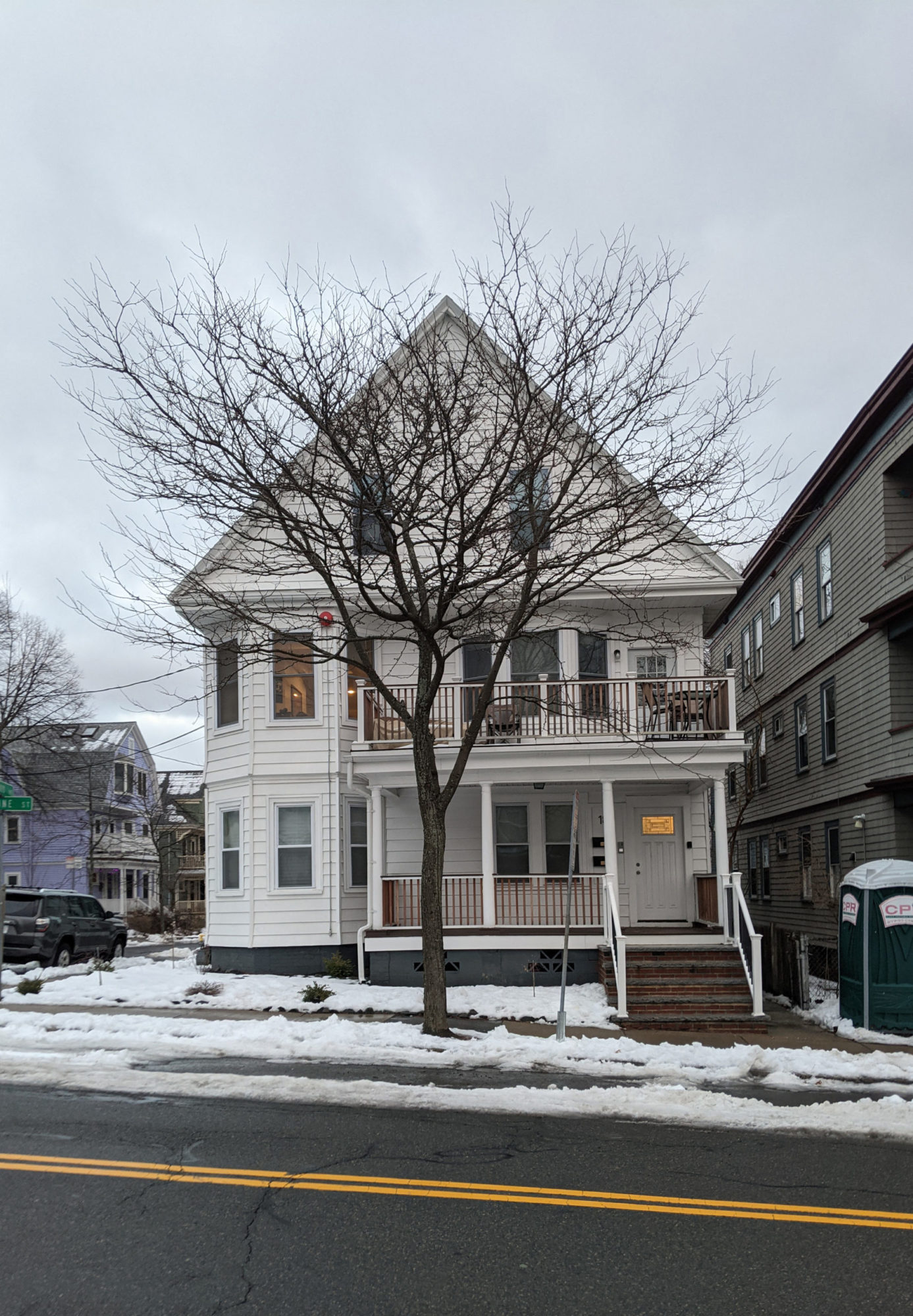 A straight on, across the street view of a multi story, white, wooden house. Stairs lead up to the front door on the front right of the house.. There are porches on the front right of the first and second floors. There are bay windows on the front left of the first and second floors. A tree is directly in front of the house. There is a port-a-potty not far from the entryway stairs.