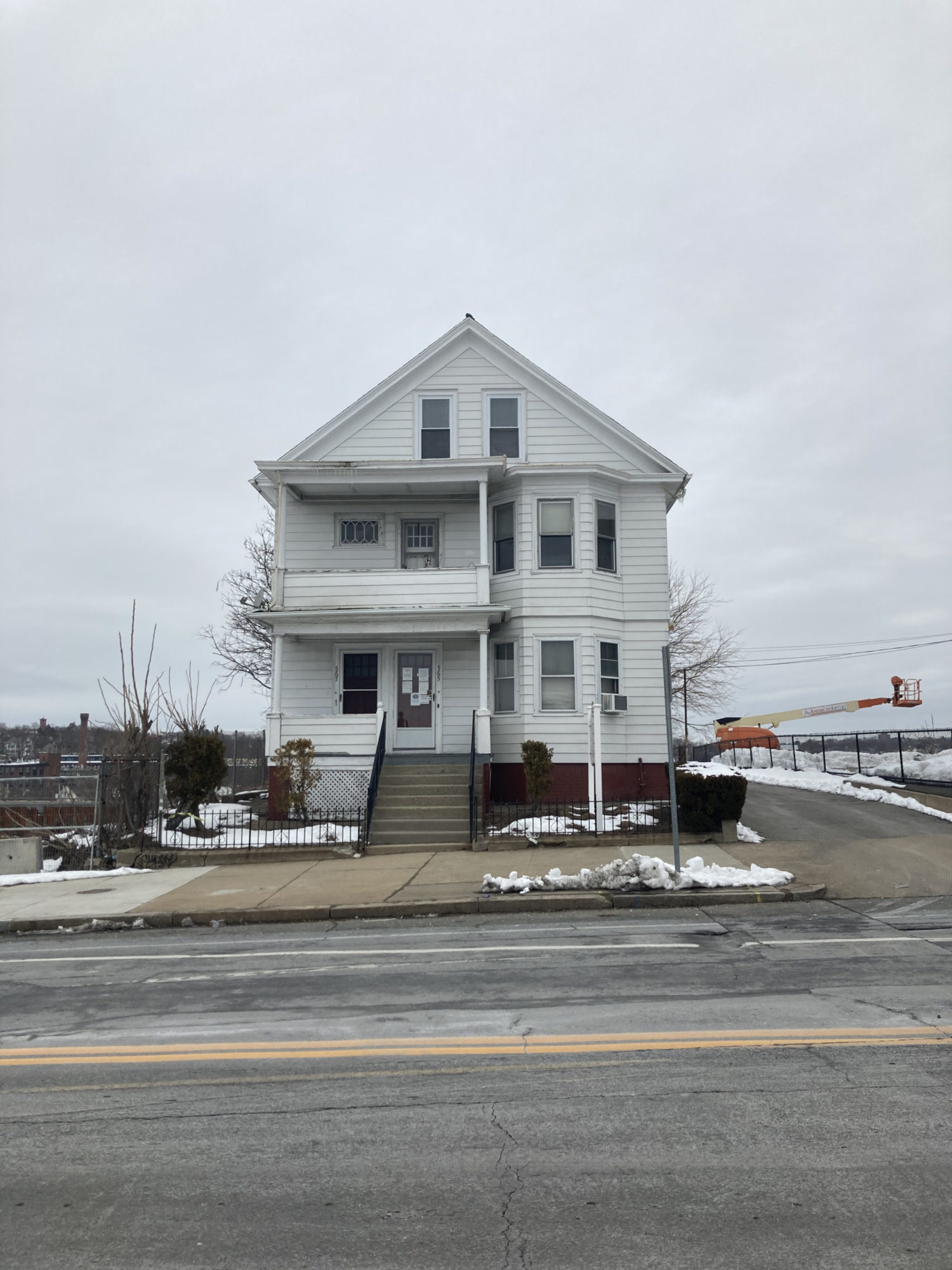 A straight on, across the street view of a muti story, white, wooden house. Stairs lead up to the front door on the front left of the building. There is are bay windows on the front right of the first and second floors. There are porches on the front left of the first and second stories. A metal fence surrounds the front yard. No other houses are in view. Construction is in view behind the house.