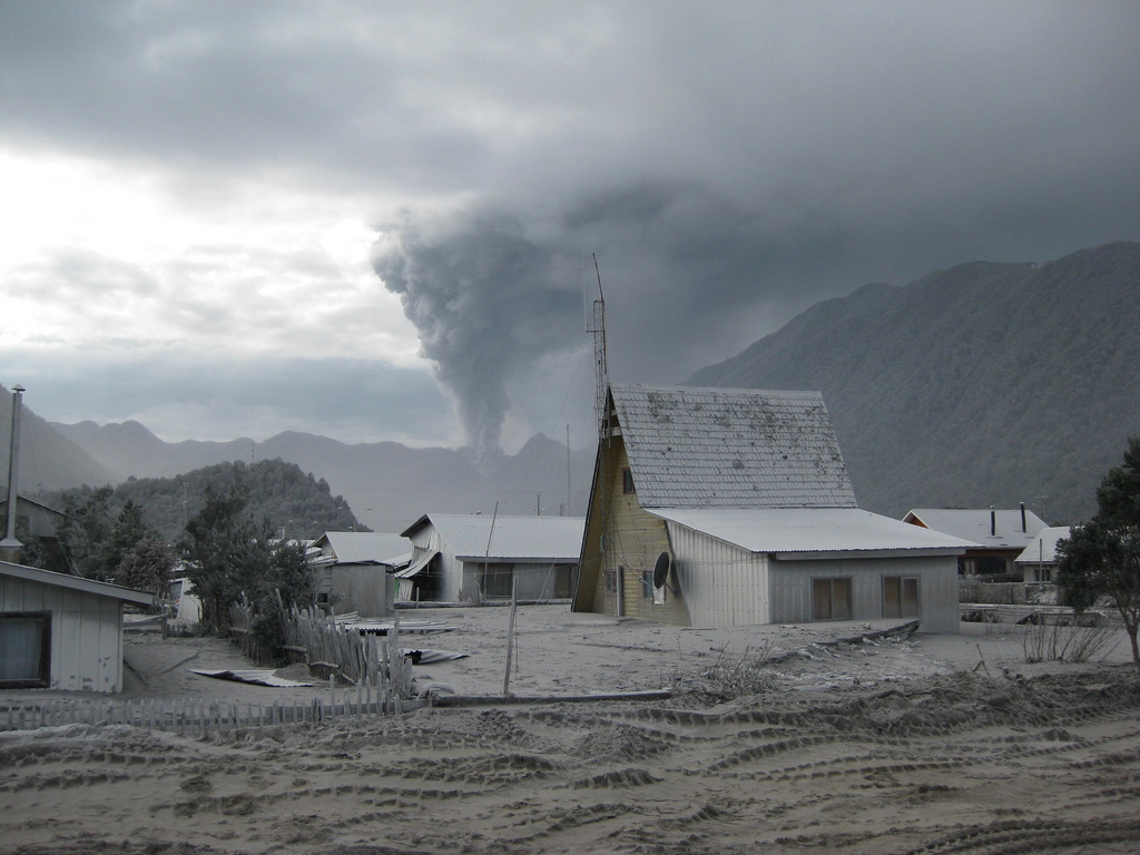 A photograph of a landscape featuring buildings in the foreground and an erupting volcano in the distant background.