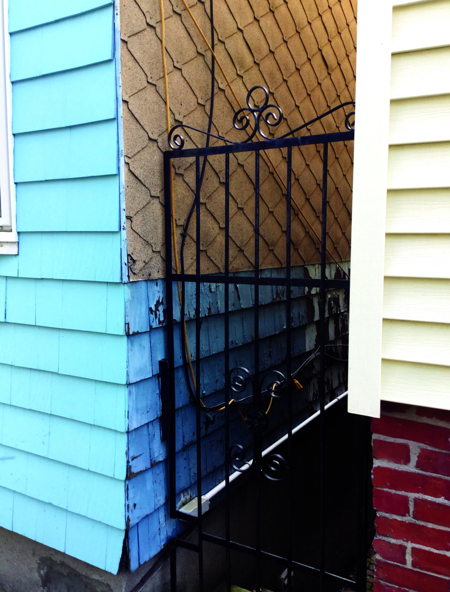A closer shot of an iron fence blocking an alley between two buildings. The building on the left is blue and brown wood and the house on the right is white wood and red brick. The fence is black. Wires go down the sides of the buildings, leading behind the fence.
