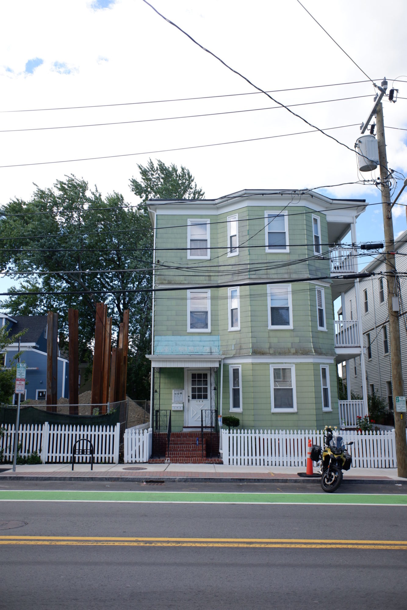 A straight on, street view image of a three story muted green house. The front door is on the front left of the building under an awning. The paint directly above the awning is much more blue than the rest of the building. There is a bay window on the front left side of all three floors. There are also covered porches on all three floors on the right side of the building. There is a white fence around the yard and a motorcycle and traffic cone on the side walk. Telephone wires cross in front of the house.